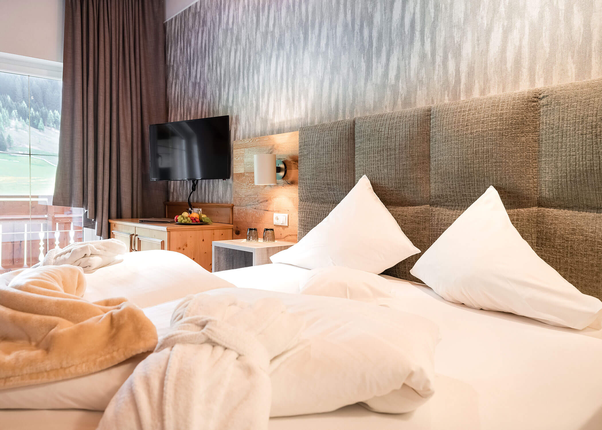 Modernes Hotelzimmer mit großem Bett, TV, Obstkorb, Bademänteln und einem Fenster mit Blick auf die Natur. - Hotel Magdalenahof