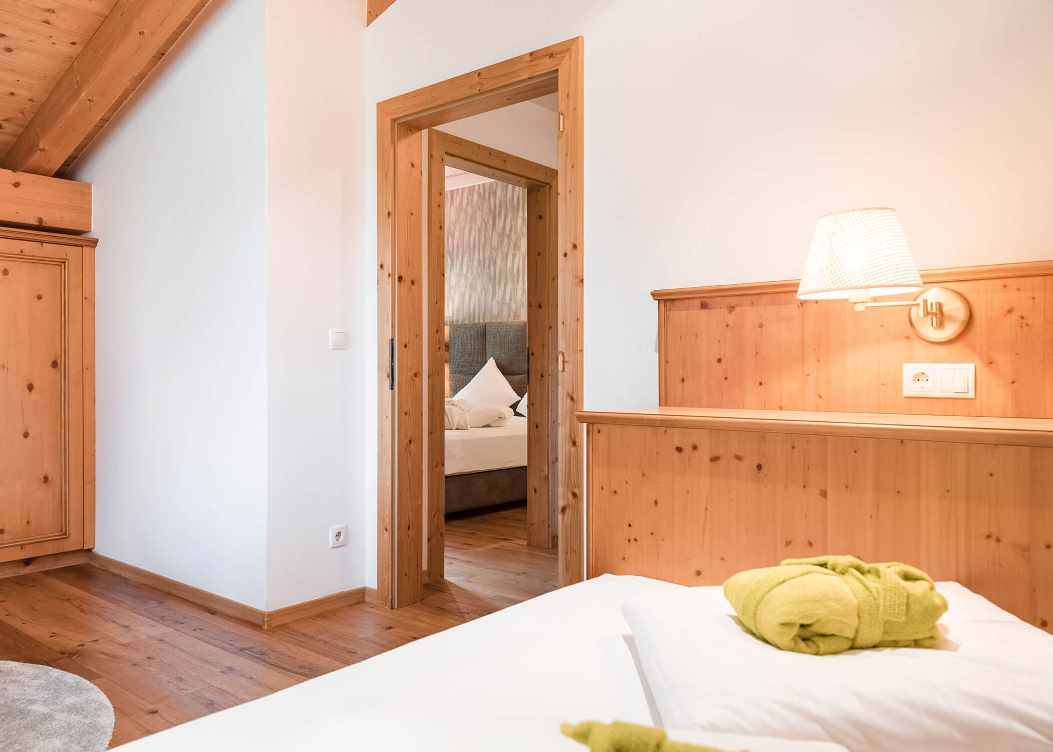 Helles Holzschlafzimmer mit weißem Bettzeug, einem Bademantel auf dem Bett und Blick in ein zweites gemütliches Schlafzimmer. - Hotel Magdalenahof