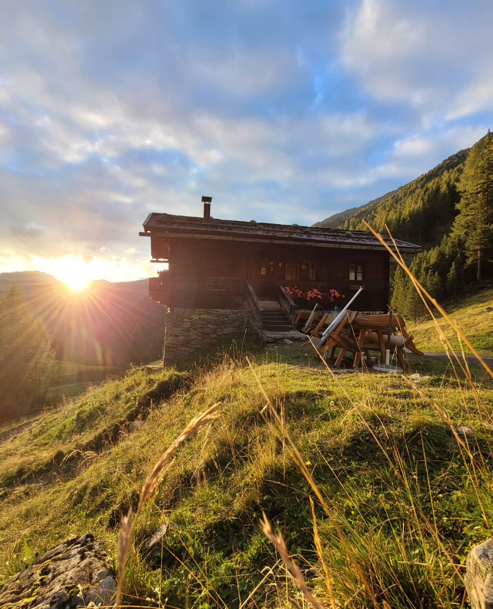 Eine Holzhütte auf einem grasbewachsenen Hügel bei Sonnenuntergang, wenn das Sonnenlicht über die entfernten Berge fällt. - Hotel Magdalenahof