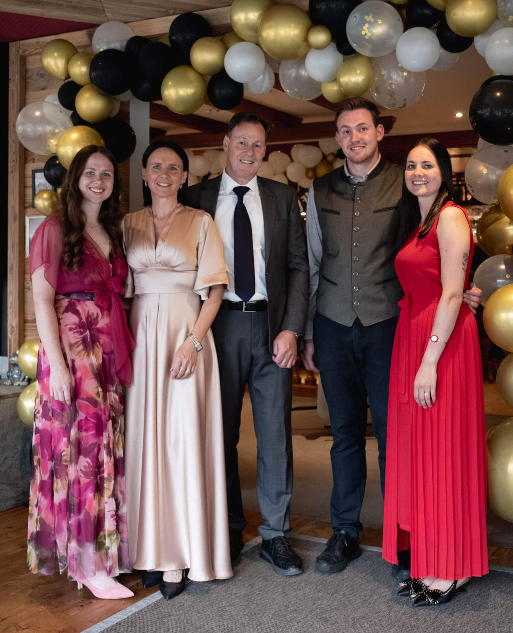 Fünf formell gekleidete Personen posieren unter einem goldenen, schwarzen und weißen Luftballonbogen bei einer Indoor-Veranstaltung. - Hotel Magdalenahof