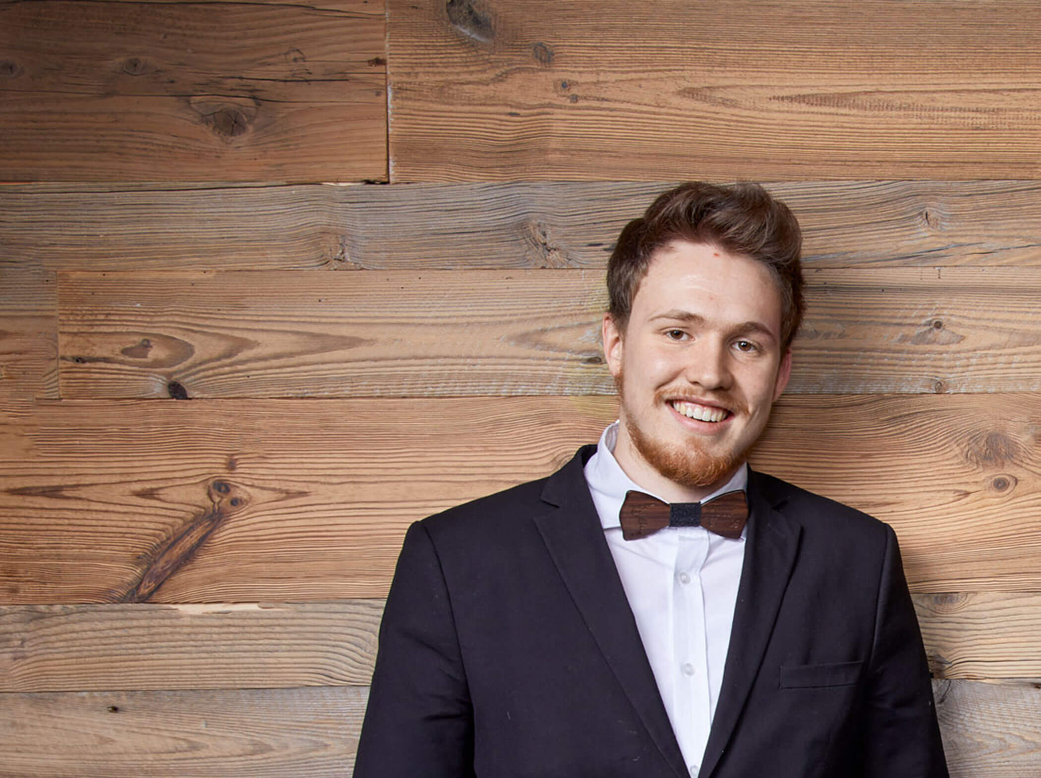 Ein lächelnder Mann in Anzug und Fliege steht vor einer Bretterwand. - Hotel Magdalenahof