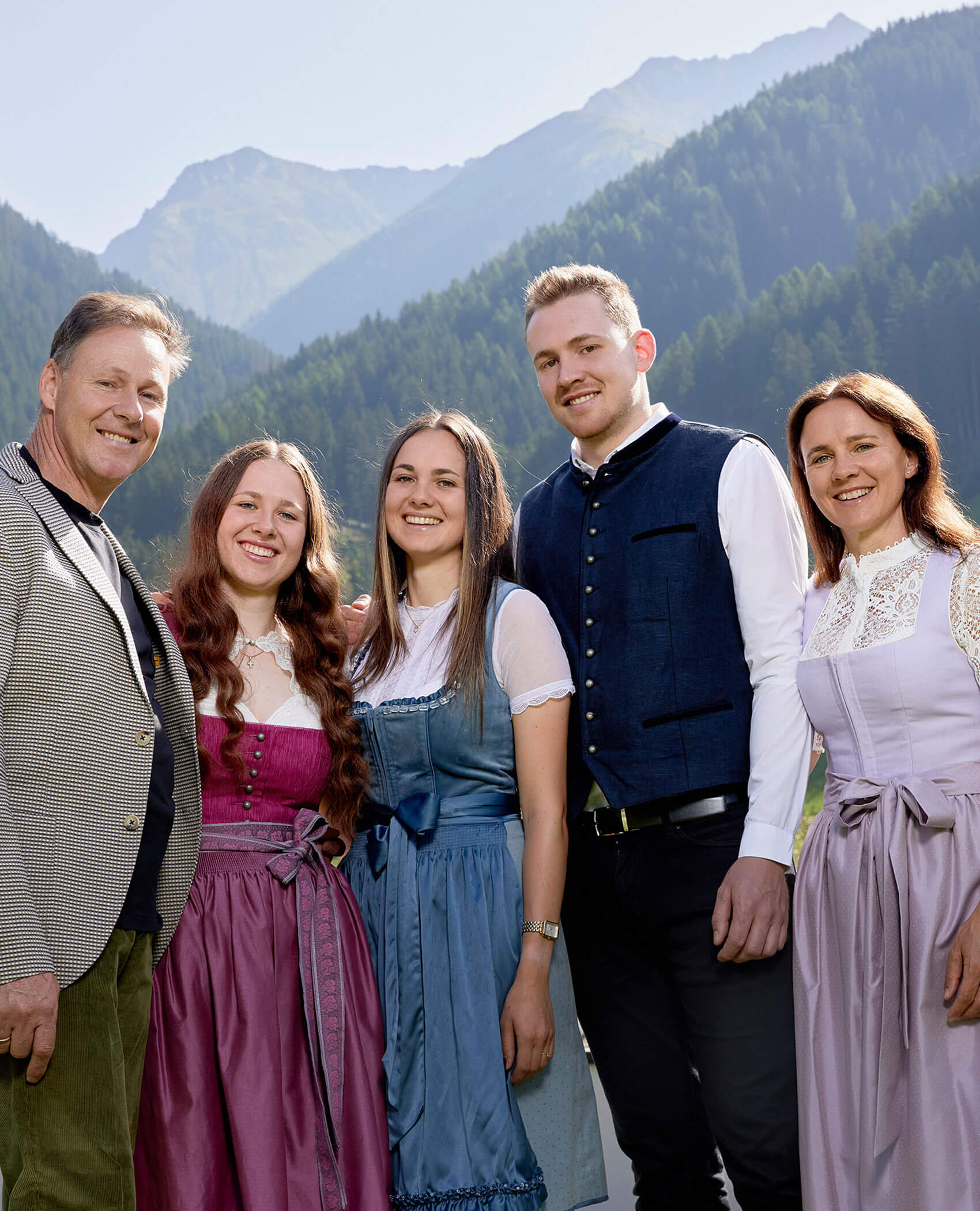 Fünf Menschen in traditioneller Kleidung stehen lächelnd im Freien, mit bewaldeten Bergen im Hintergrund. - Hotel Magdalenahof