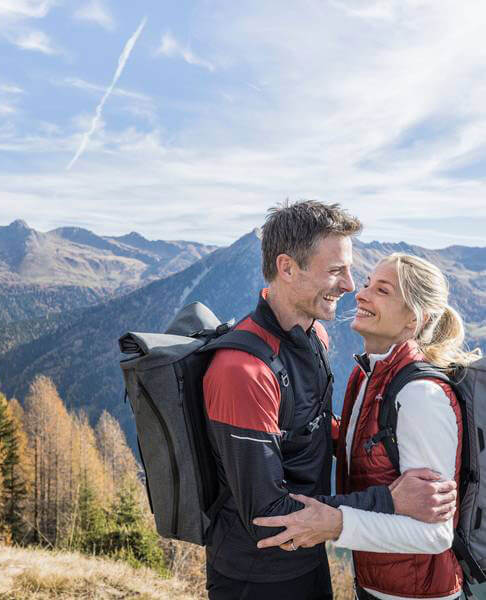 Lächelndes Paar mit Rucksäcken, das sich im Freien umarmt, Berge und blauer Himmel im Hintergrund. - Hotel Magdalenahof