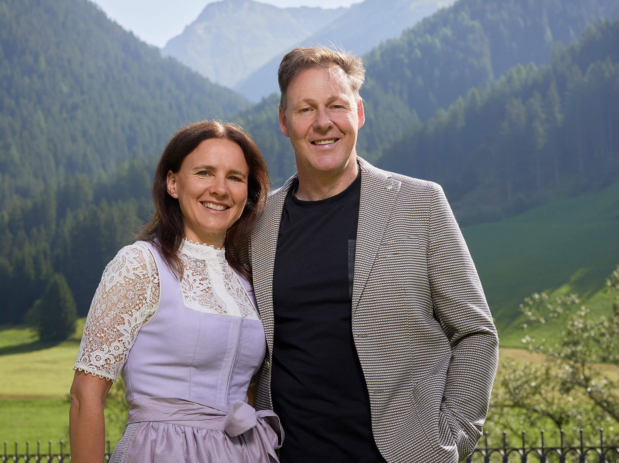 Ein lächelnder Mann und eine lächelnde Frau stehen an einem sonnigen Tag im Freien mit Bergen und Kiefern im Hintergrund. - Hotel Magdalenahof