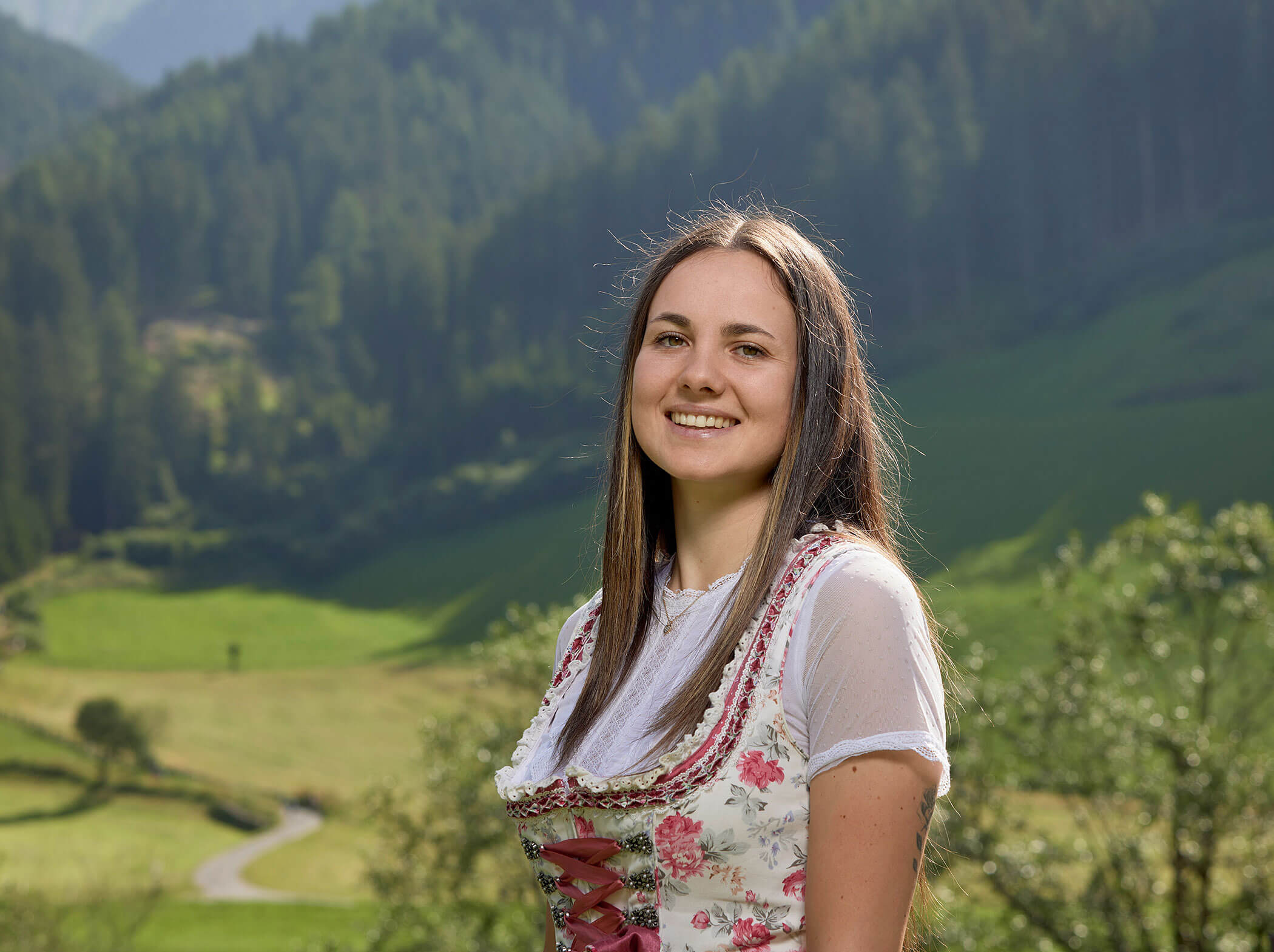 Lächelnde Frau in traditioneller Kleidung steht im Freien mit grünen Hügeln und Wald im Hintergrund. - Hotel Magdalenahof