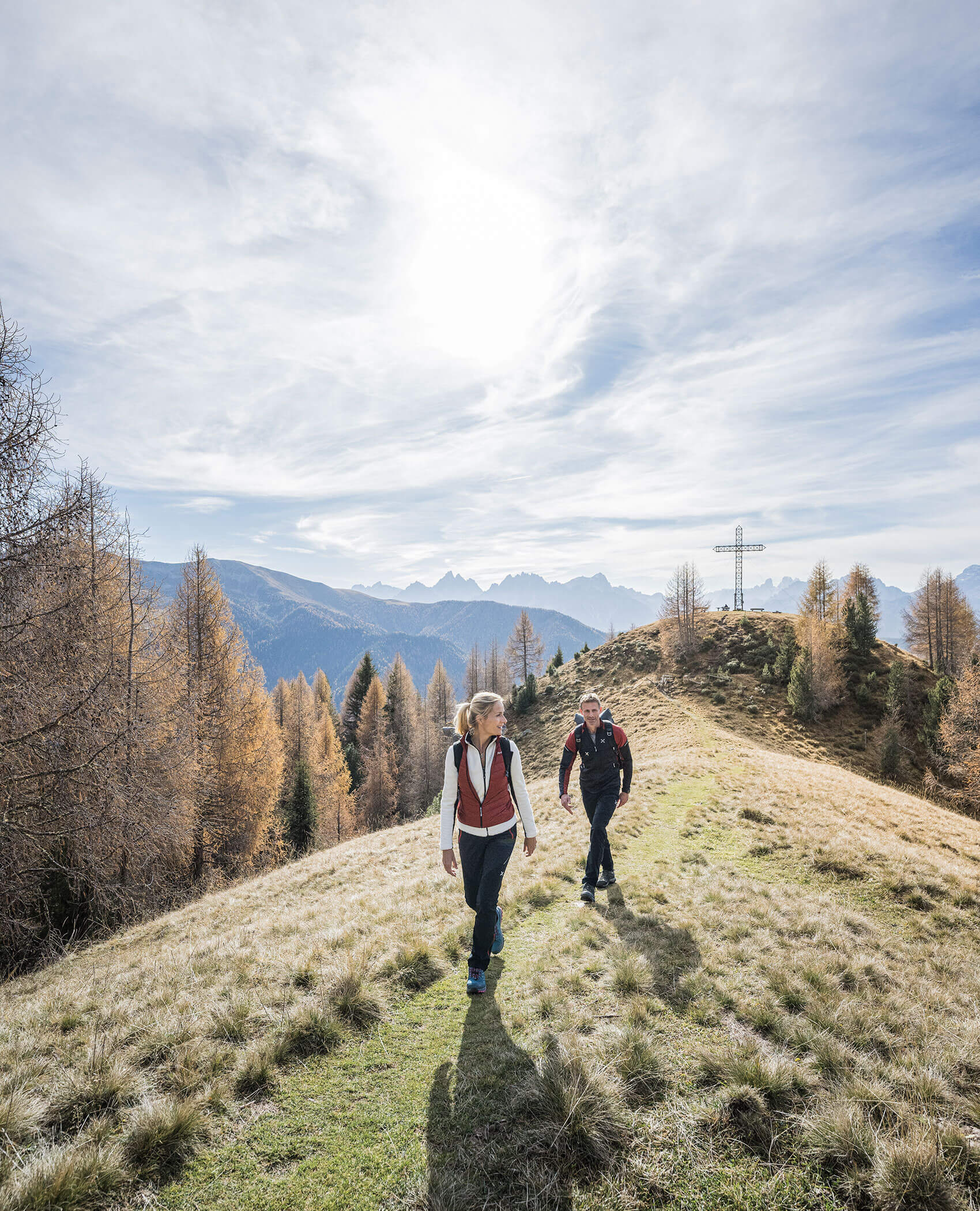 Zwei Personen wandern auf einen grasbewachsenen Hügel mit Bergen und einem großen Kreuz im Hintergrund unter einem hellen Himmel. - Hotel Magdalenahof
