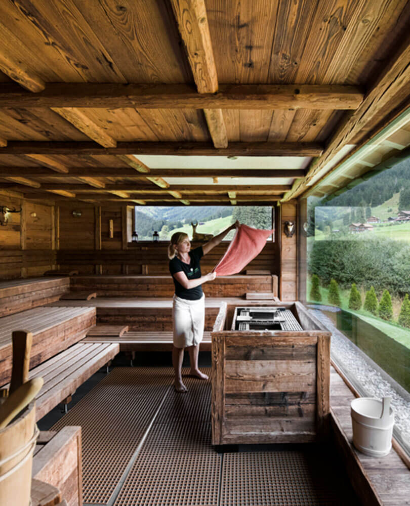 Eine Frau in einer Holzsauna schwenkt ein Handtuch vor einem großen Fenster mit Blick auf einen grünen Hügel. - Hotel Magdalenahof