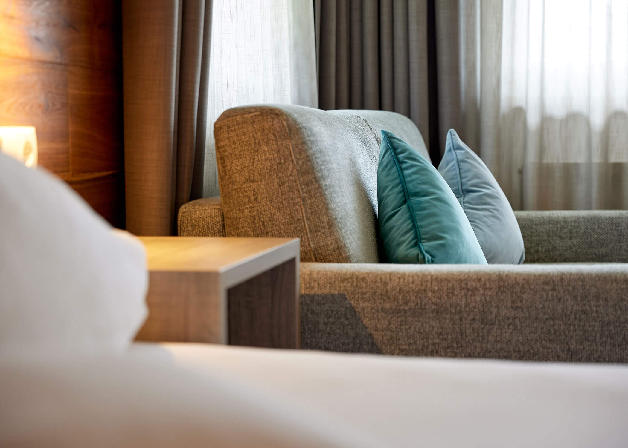 Ein grauer Sessel mit blaugrünen Kissen neben einem hölzernen Nachttisch in einem sanft beleuchteten Raum mit Vorhängen. - Hotel Magdalenahof