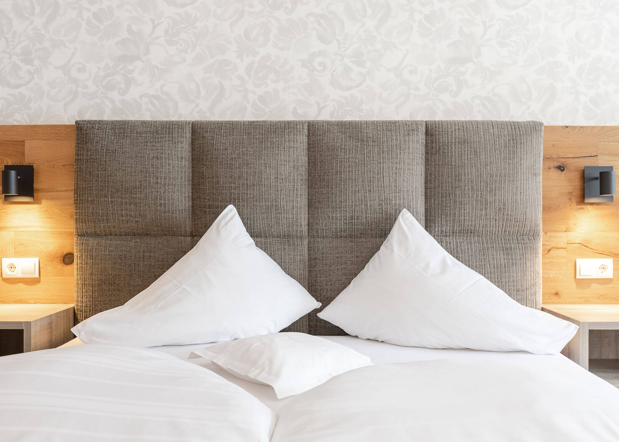 Ein ordentlich gemachtes Bett mit zwei großen Kissen vor einem gepolsterten Kopfteil und Wandleuchten auf jeder Seite. - Hotel Magdalenahof