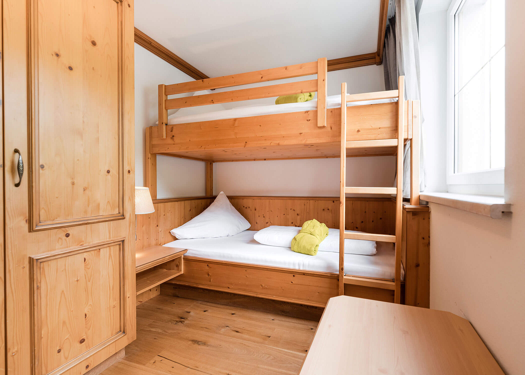 Kleines Schlafzimmer mit Etagenbetten aus Holz, einem Kleiderschrank und einem Fenster, das Tageslicht hereinlässt. - Hotel Magdalenahof