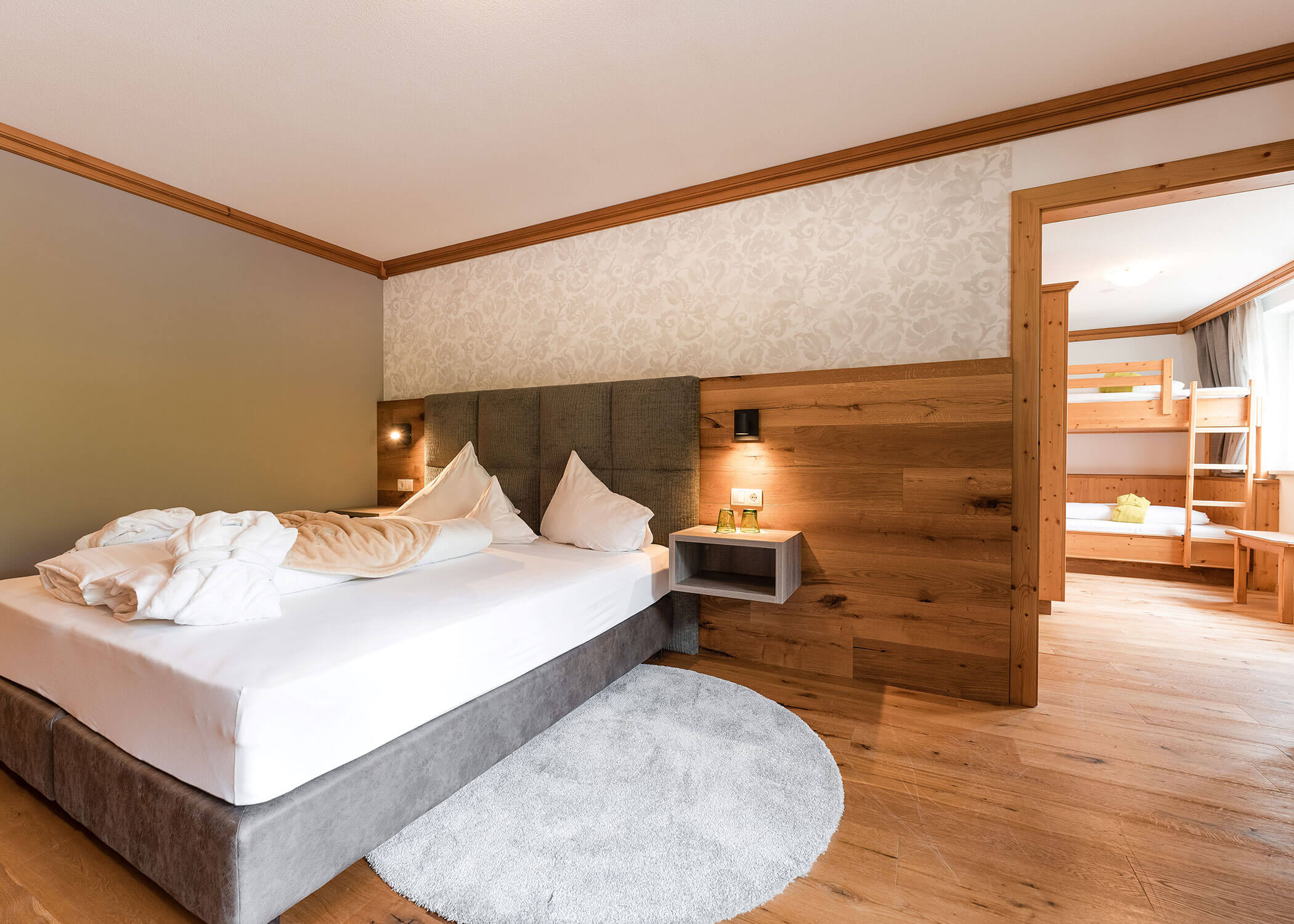 Modernes Hotelzimmer mit einem Doppelbett, sanfter Beleuchtung, Holzakzenten und einem Nebenzimmer mit Etagenbetten. - Hotel Magdalenahof