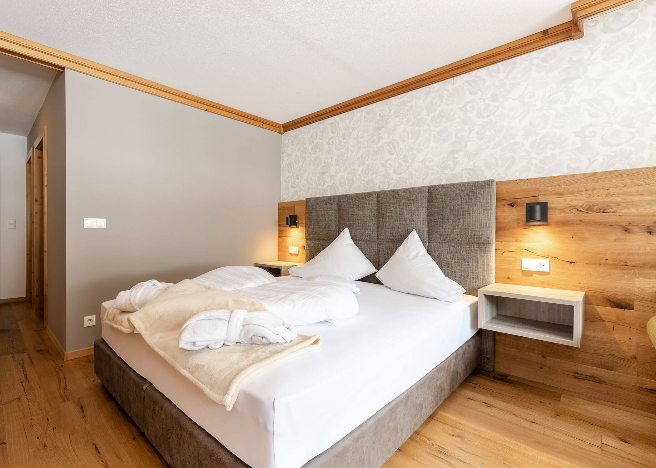 Modernes Hotelzimmer mit einem Doppelbett, weißer Bettwäsche, zwei Bademänteln und Holzakzenten an Wänden und Boden. - Hotel Magdalenahof