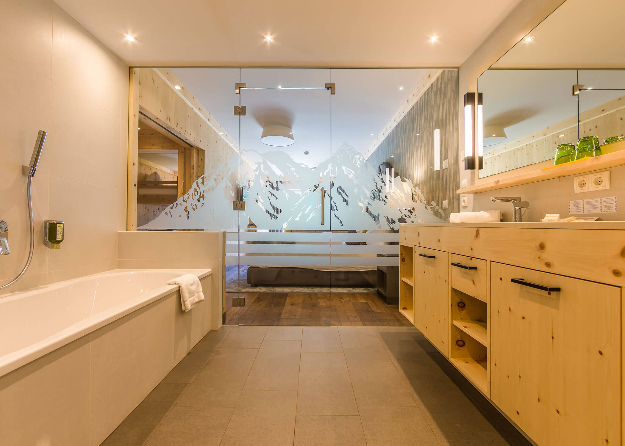 Modernes Badezimmer mit Holzschränken, einer Badewanne und einer Milchglaswand mit Bergmotiven. - Hotel Magdalenahof
