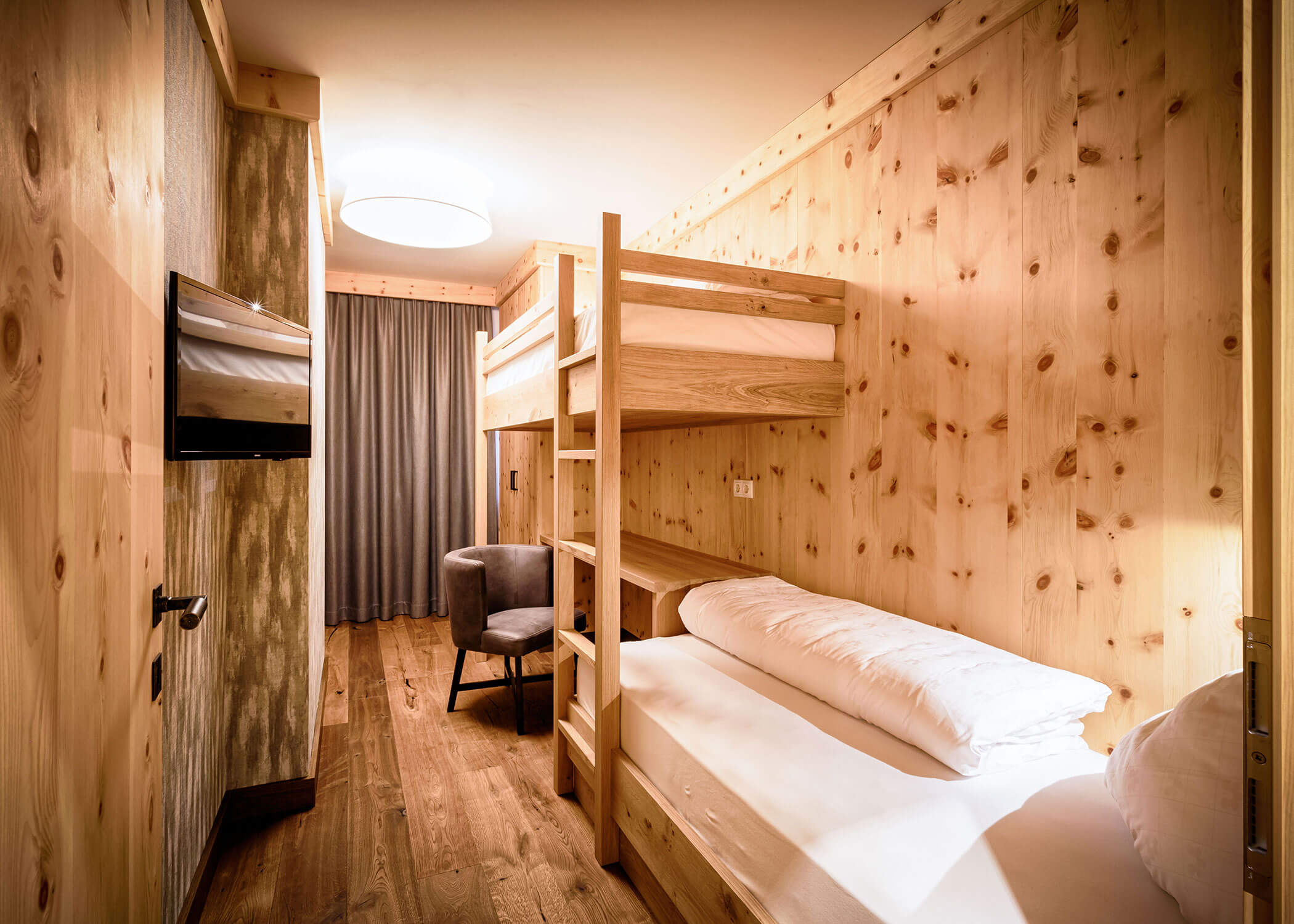 Gemütliches Holzzimmer mit Etagenbetten, einem an der Wand montierten Fernseher, einem Stuhl und raumhohen Vorhängen. - Hotel Magdalenahof