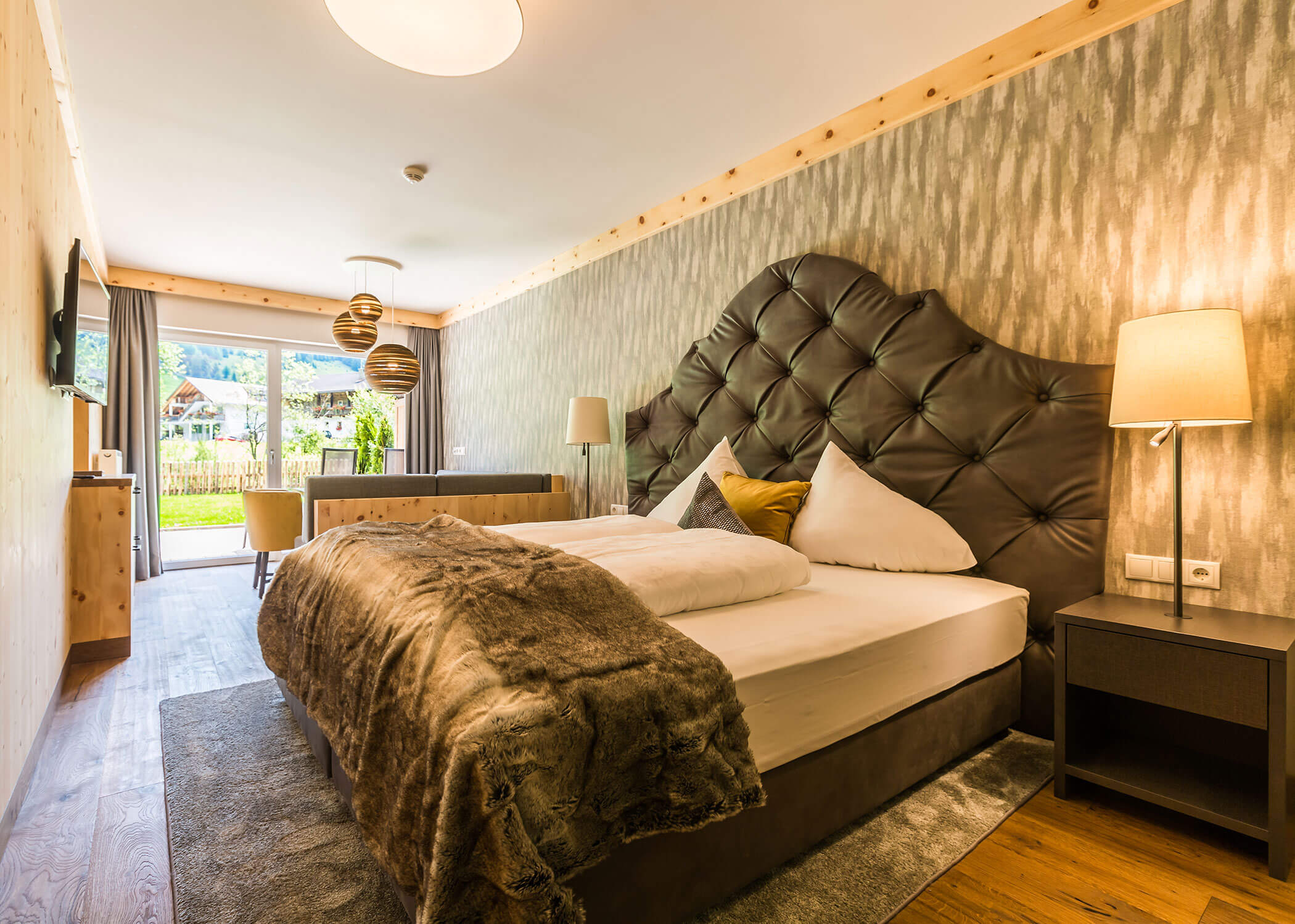 Modernes Hotelzimmer mit großem Bett, Felldecke, Lampen und Blick aus dem Fenster auf einen Garten und die Berge. - Hotel Magdalenahof