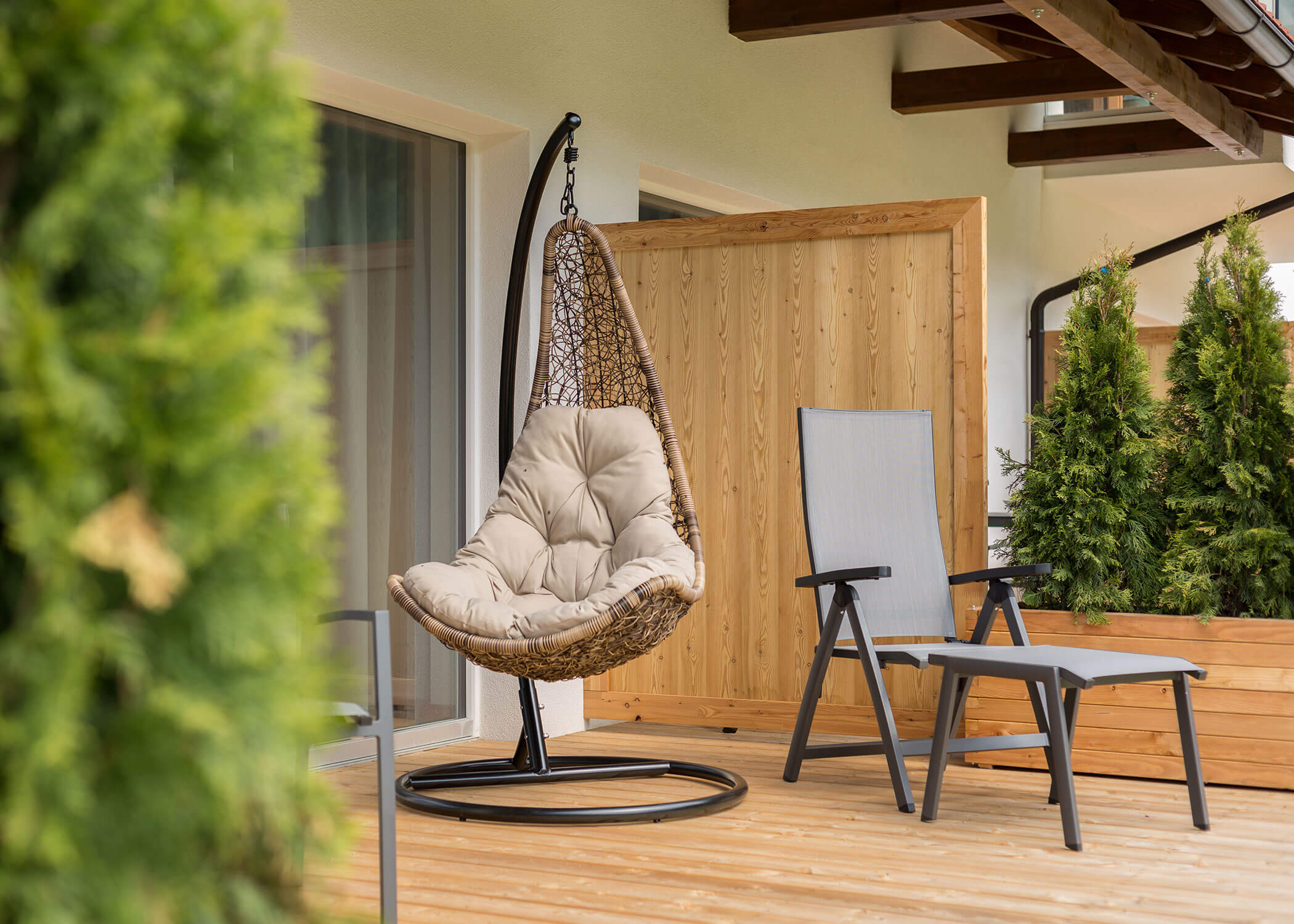 Ein Hängekorb und ein Loungesessel mit einem Tisch auf einer Holzterrasse, umgeben von Grünpflanzen. - Hotel Magdalenahof