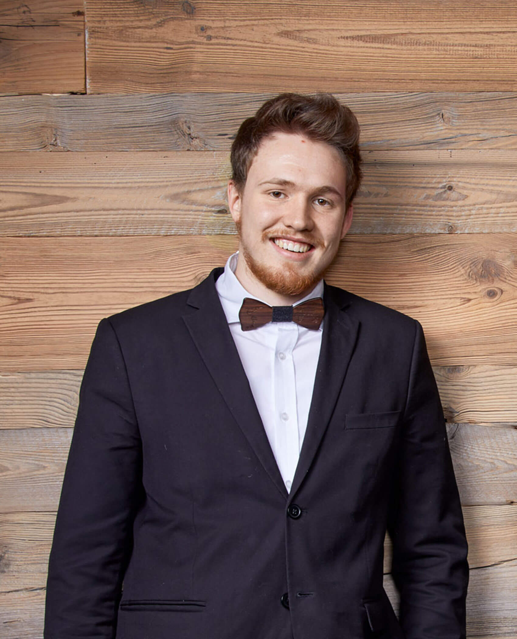 Ein lächelnder Mann in Anzug und Fliege steht vor einer Holzwand. - Hotel Magdalenahof