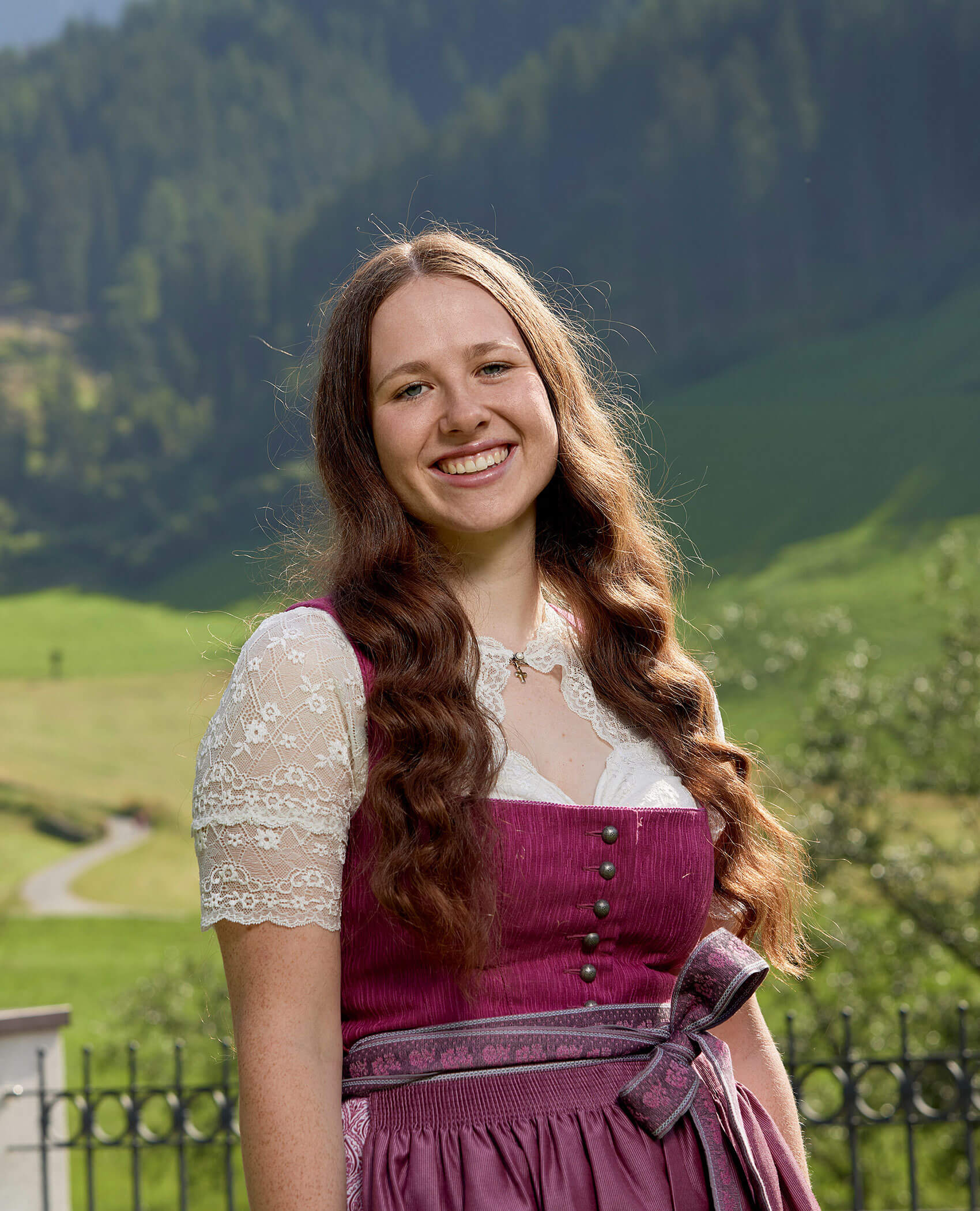 Lächelnde junge Frau in traditioneller Kleidung steht im Freien mit grünen Hügeln und Wald im Hintergrund. - Hotel Magdalenahof
