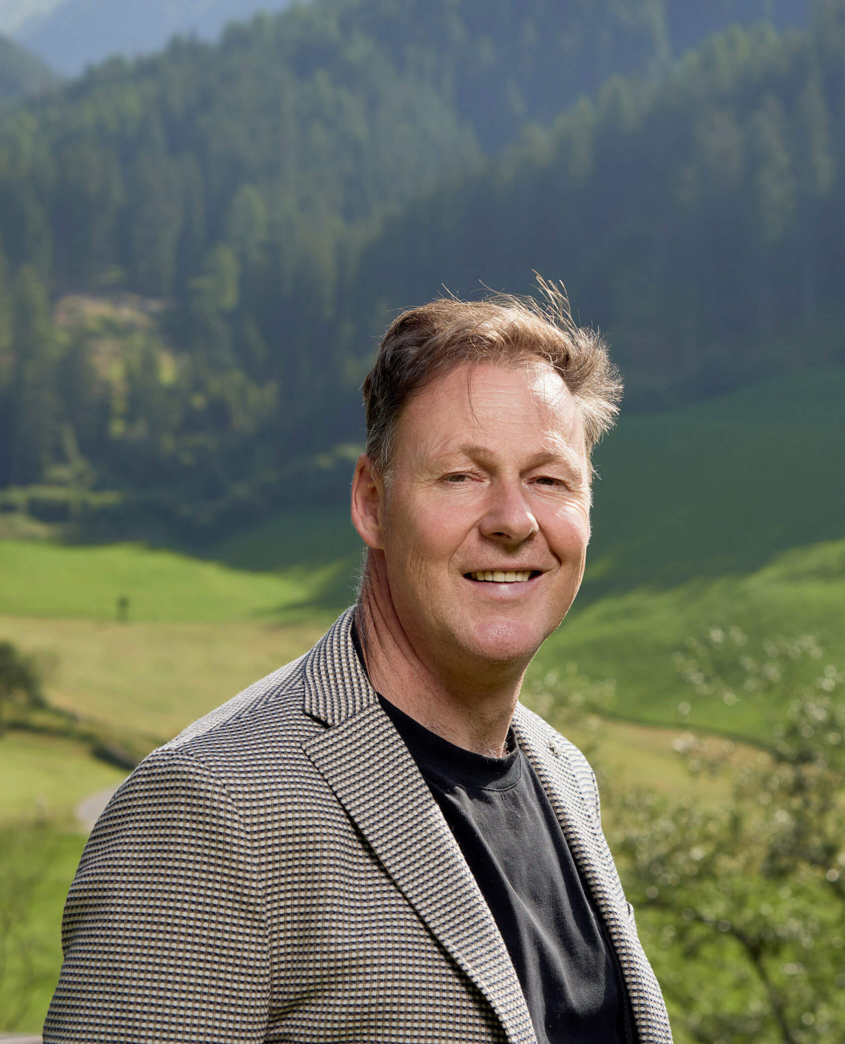 Ein lächelnder Mann in einem karierten Blazer steht im Freien mit grünen Hügeln und Bäumen im Hintergrund. - Hotel Magdalenahof
