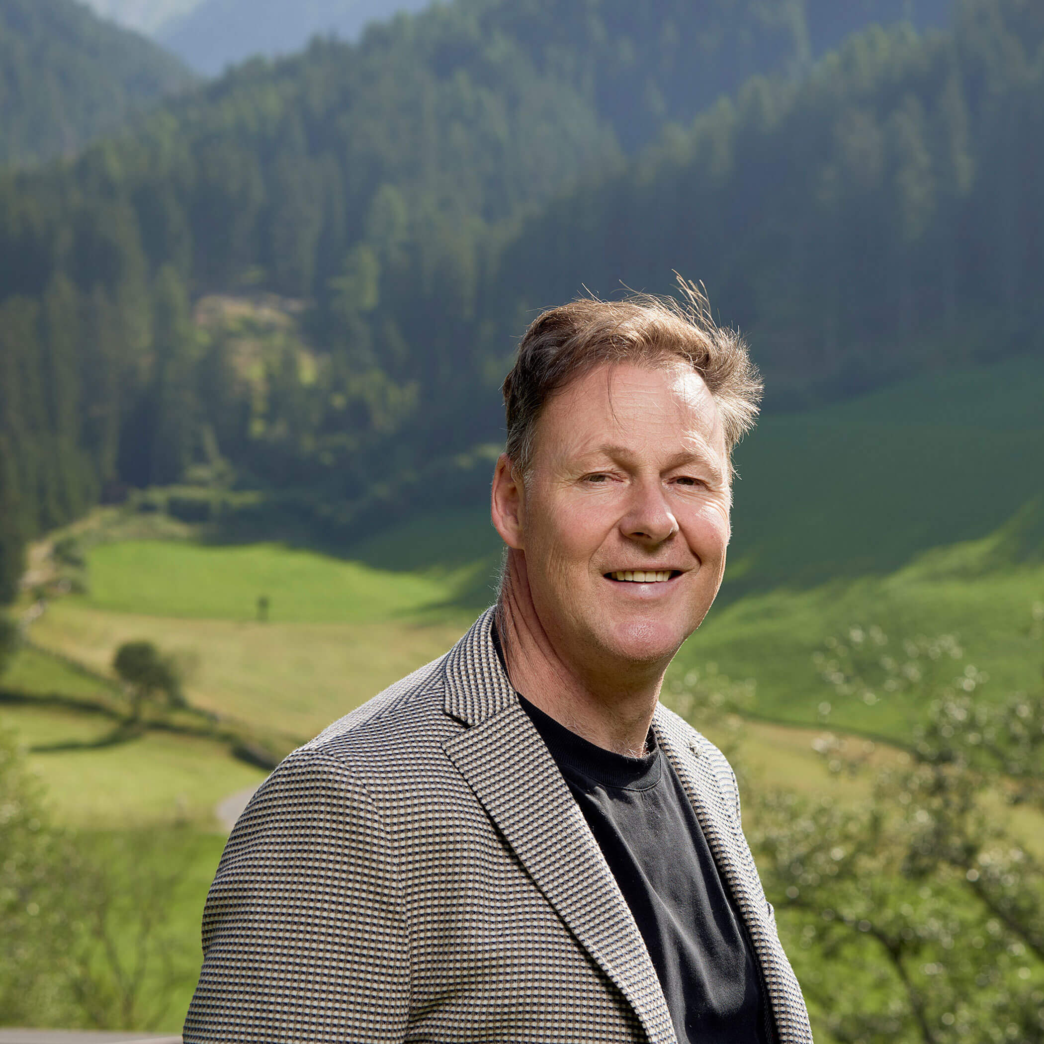 Ein lächelnder Mann in einer karierten Jacke steht im Freien mit grünen Hügeln und Bäumen im Hintergrund. - Hotel Magdalenahof