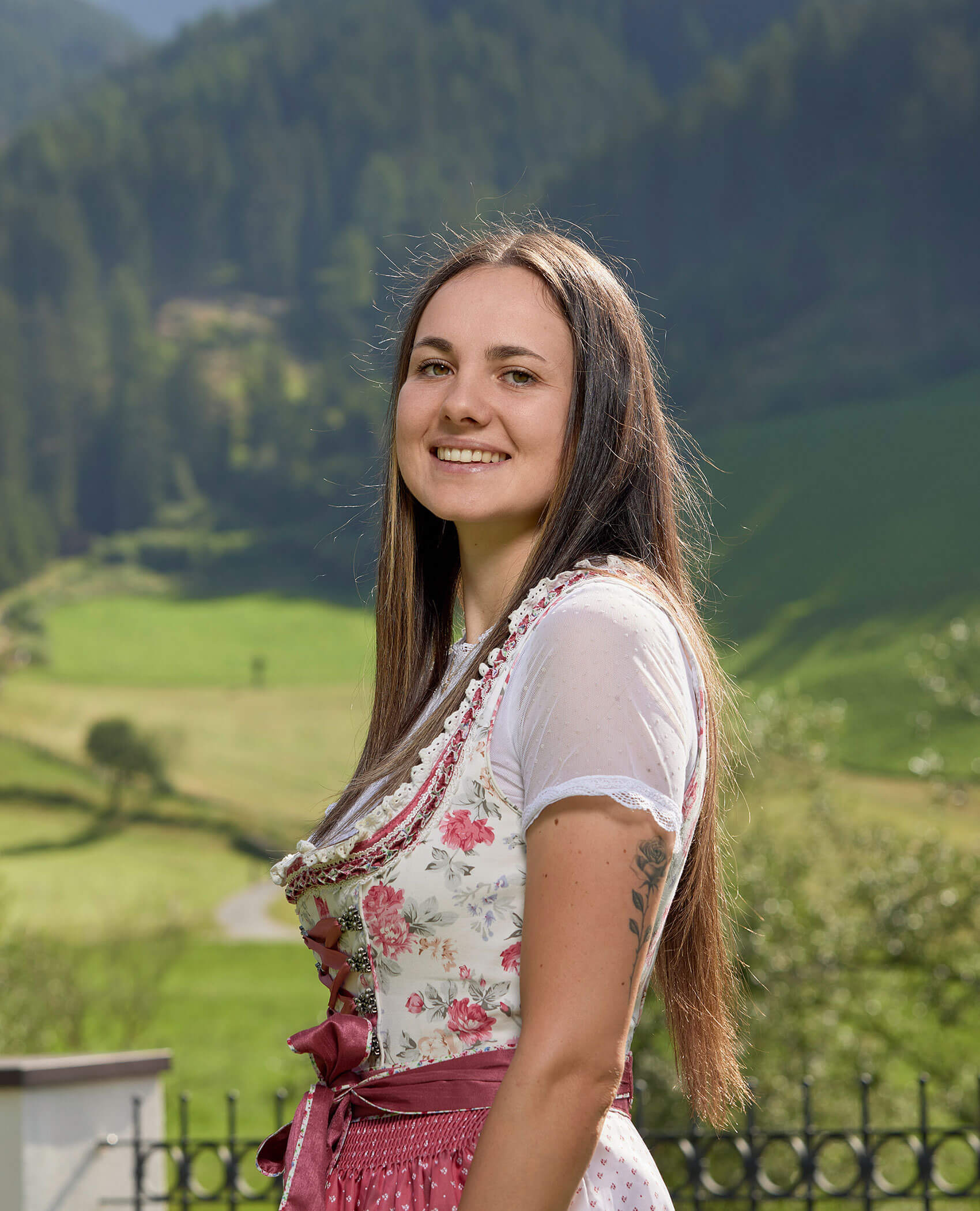 Eine lächelnde Frau in einem geblümten Dirndl steht im Freien mit grünen Hügeln und Bäumen im Hintergrund. - Hotel Magdalenahof
