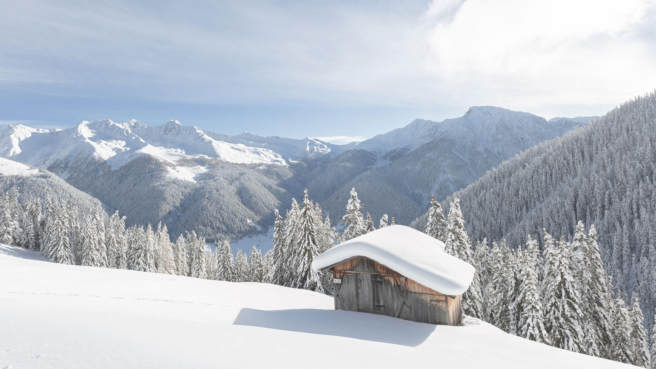 A snow-covered cabin sits among snowy trees with mountains in the background under a bright sky. - Hotel Magdalenahof