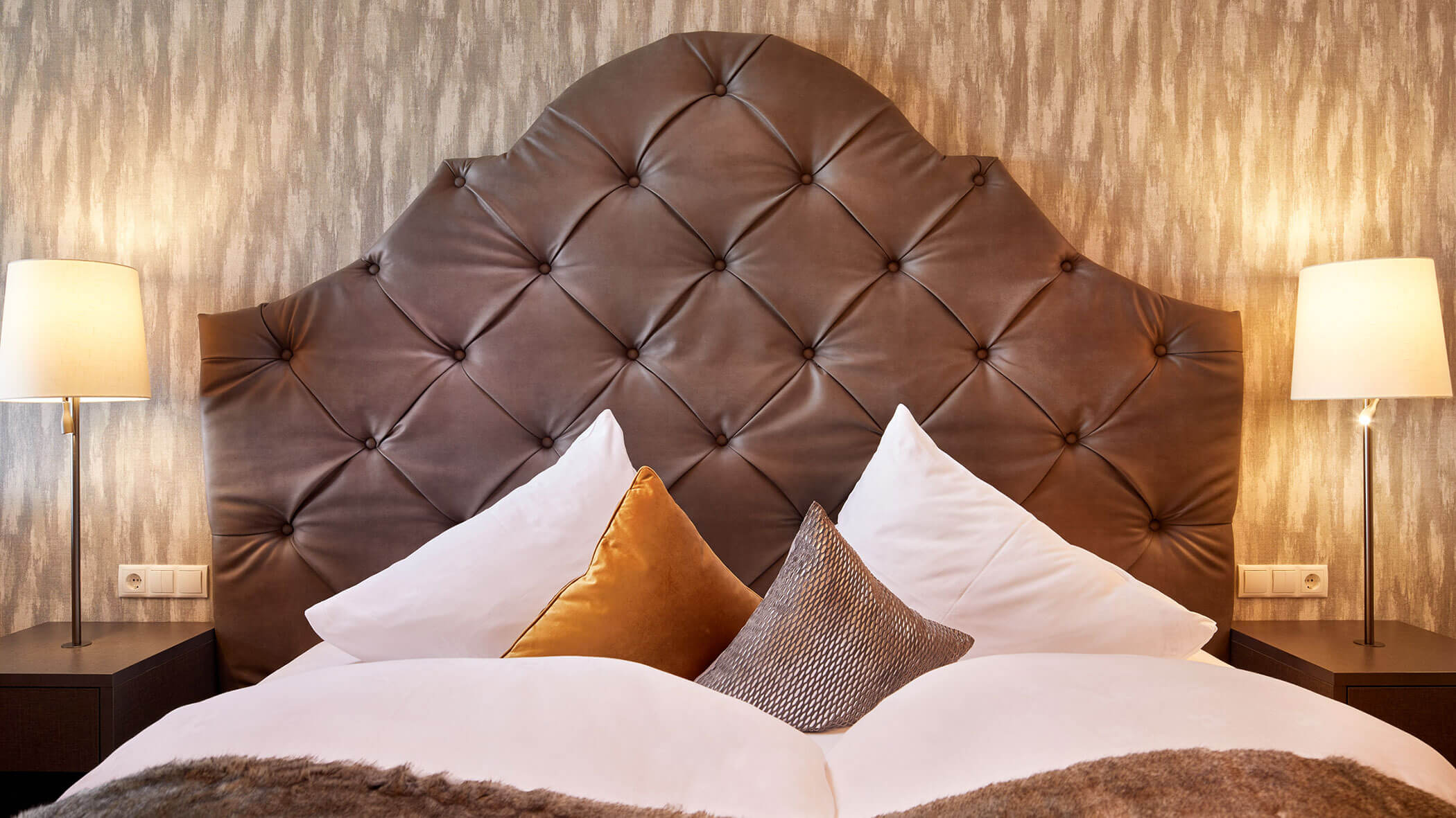Luxurious bed with a tufted leather headboard, plush pillows, and bedside lamps on each side. - Hotel Magdalenahof