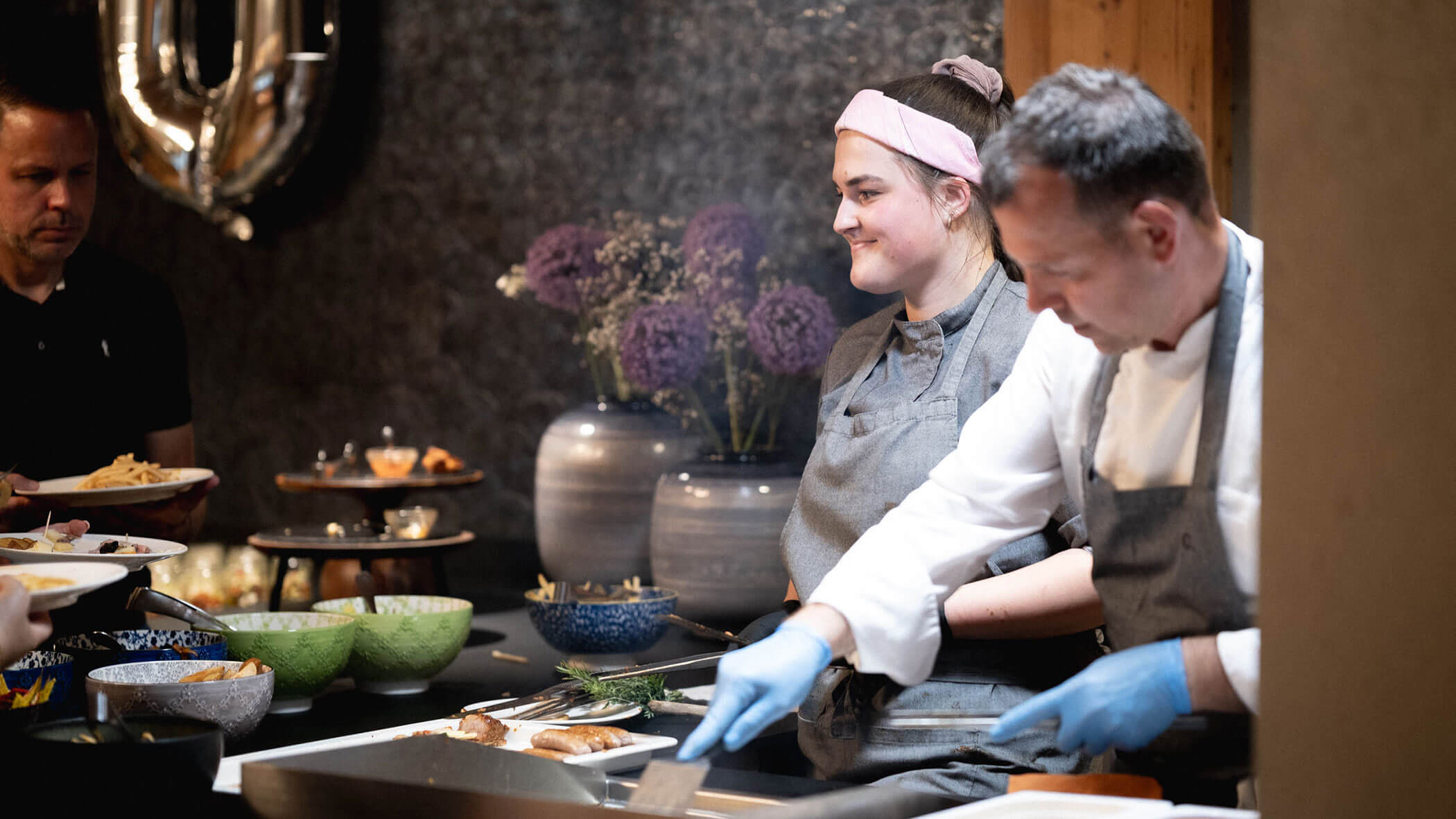 Two chefs cook and plate food in a kitchen, with bowls and vases of purple flowers in the background. - Hotel Magdalenahof