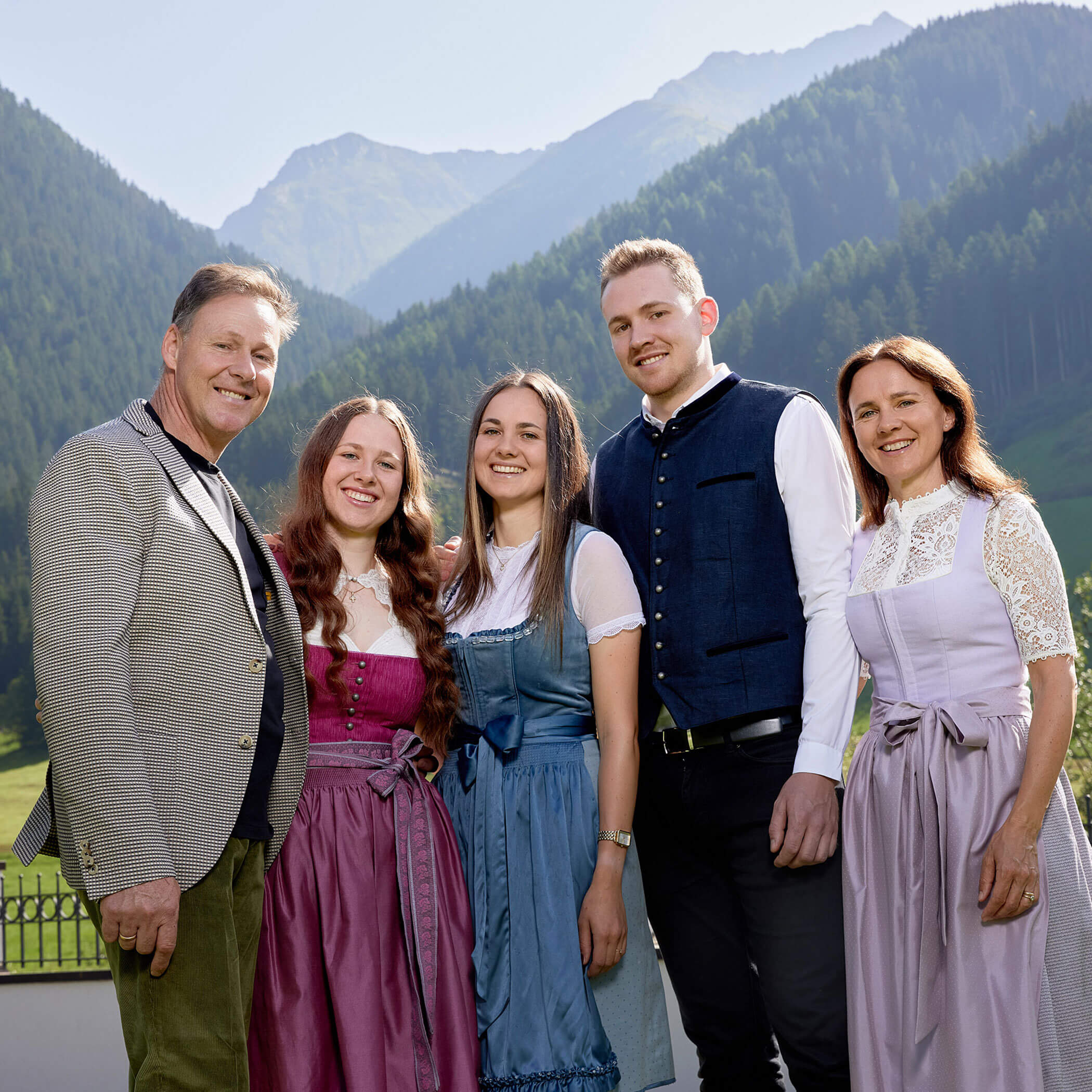A family of five in traditional clothing poses outdoors with mountains and trees in the background. - Hotel Magdalenahof