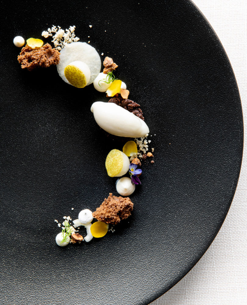 Kunstvoll angerichtetes modernes Dessert mit Klecksen, Streuseln und Garnierungen auf einem schwarzen Teller in einer geschwungenen Linie. - Hotel Magdalenahof