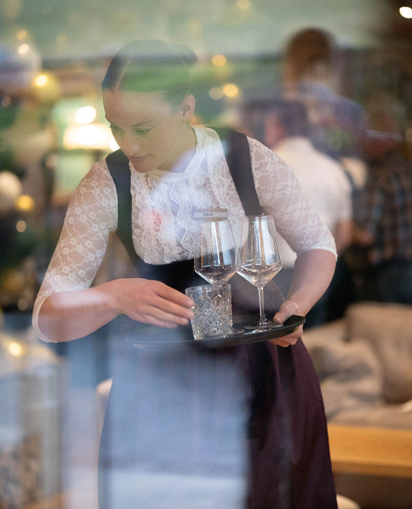 Eine Kellnerin in weißem Spitzenoberteil und Schürze trägt in einem Restaurant ein Tablett mit Gläsern. - Hotel Magdalenahof