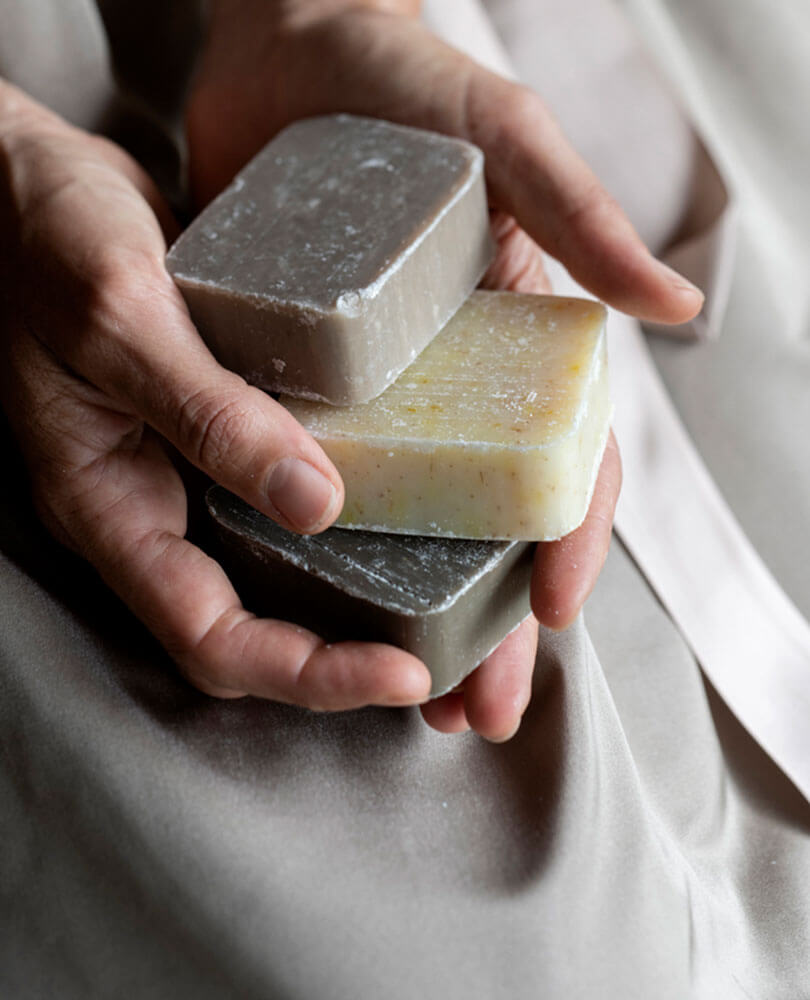 Die Hände halten drei rechteckige Seifenstücke in verschiedenen Farben auf einem weichen, neutralen Stoffhintergrund. - Hotel Magdalenahof
