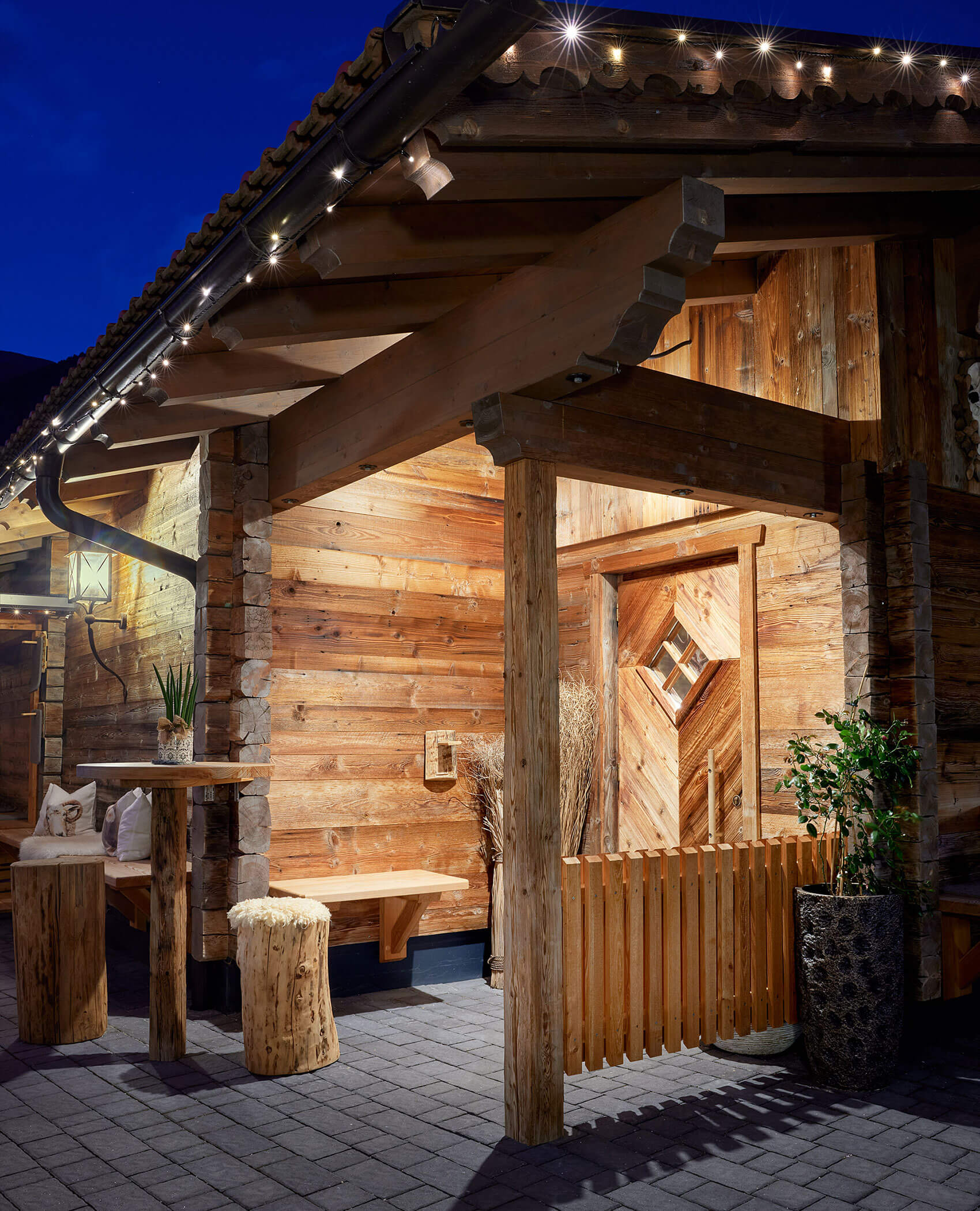Rustikale Holzhütte bei Nacht, warm beleuchtet mit Lichterketten und Möbeln im Blockhausstil auf einer Steinterrasse. - Hotel Magdalenahof