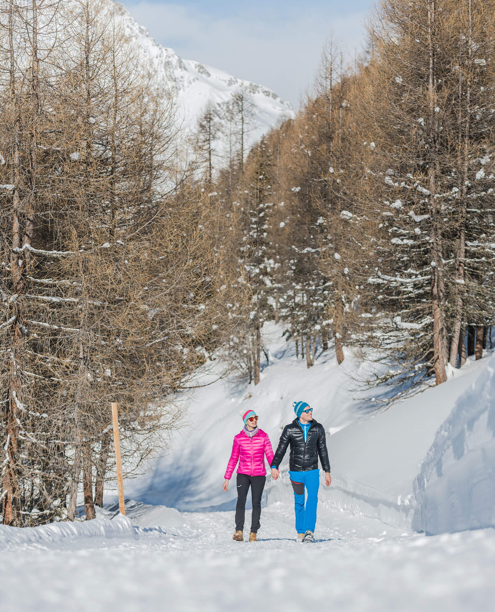 Zwei Personen in Winterkleidung gehen Hand in Hand auf einem verschneiten Weg durch ein bewaldetes Berggebiet. - Hotel Magdalenahof