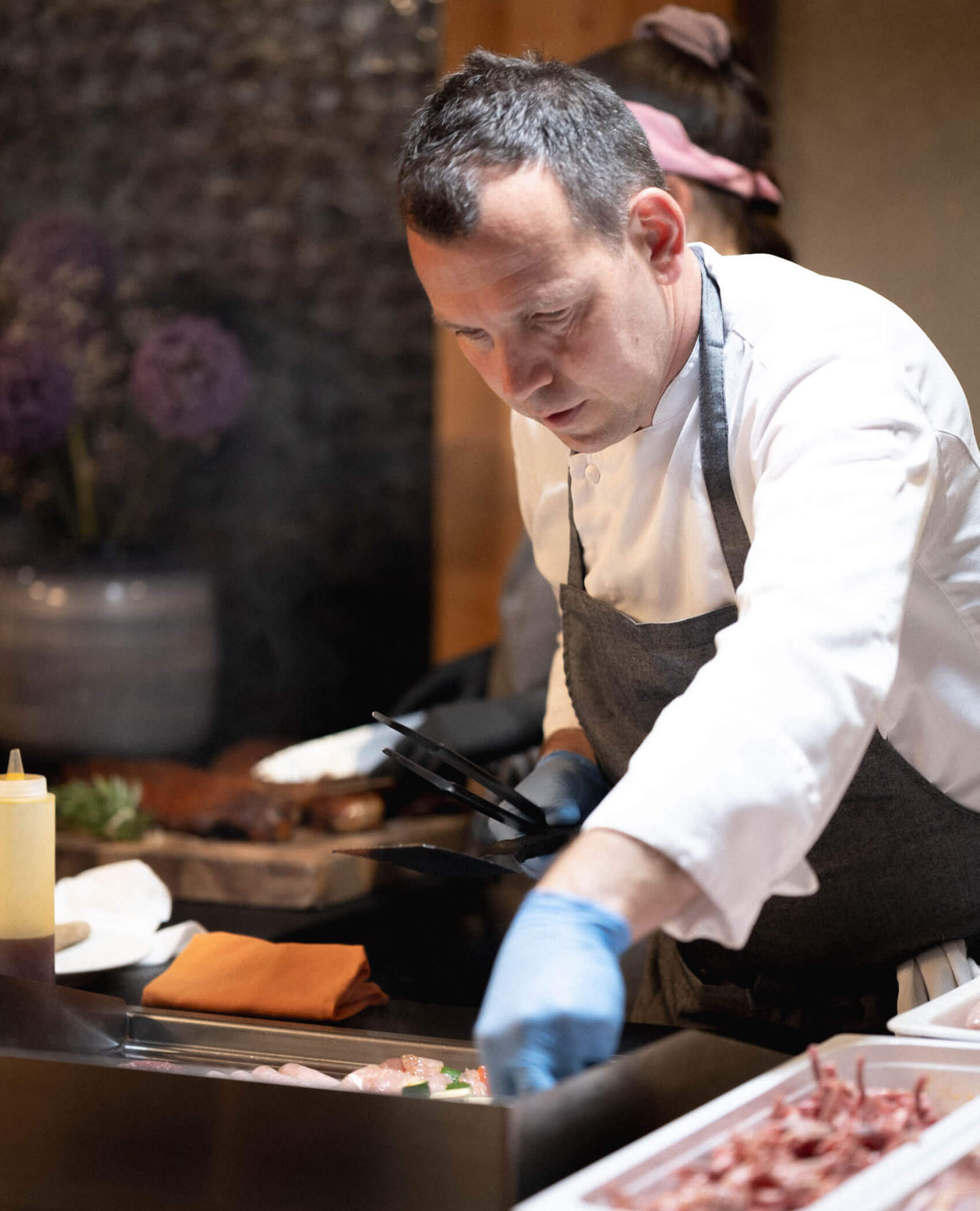 Ein Koch in weißem Kittel und Schürze bereitet an einem Tresen das Essen zu, konzentriert auf seine Arbeit. - Hotel Magdalenahof