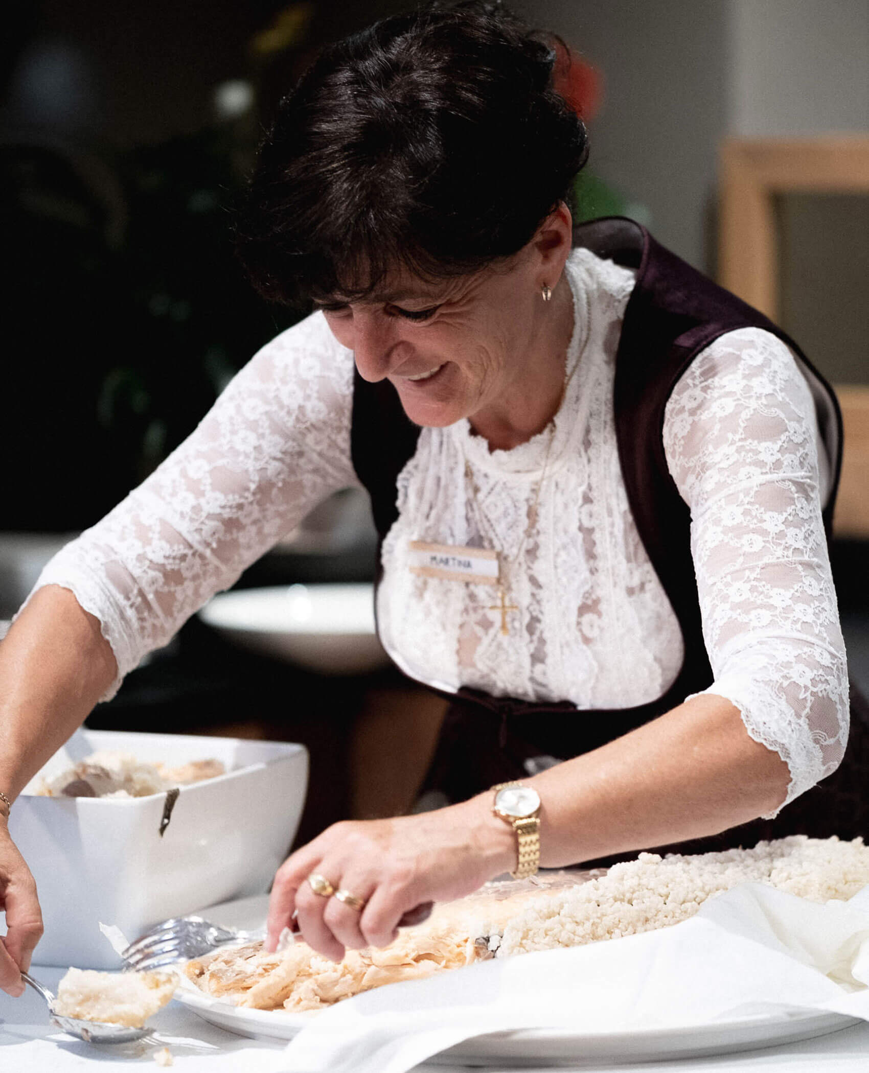 Eine lächelnde Frau in einem Spitzenoberteil bereitet das Essen an einem Tisch vor, wobei sie die Stücke mit Gabel und Löffel anrichtet. - Hotel Magdalenahof