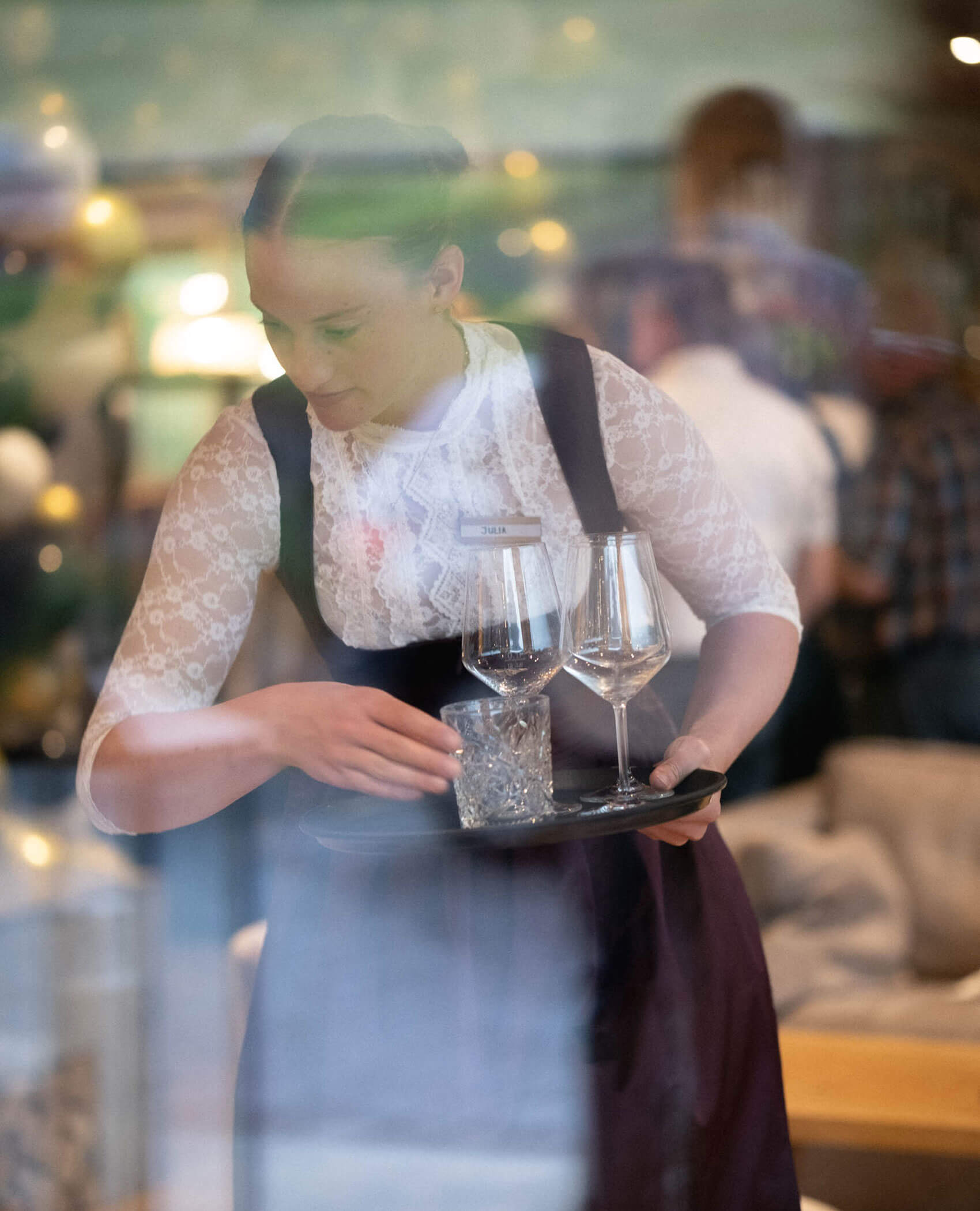 Eine Kellnerin in weißer Spitzenbluse ordnet Gläser auf einem Tablett in einem Restaurant an. - Hotel Magdalenahof
