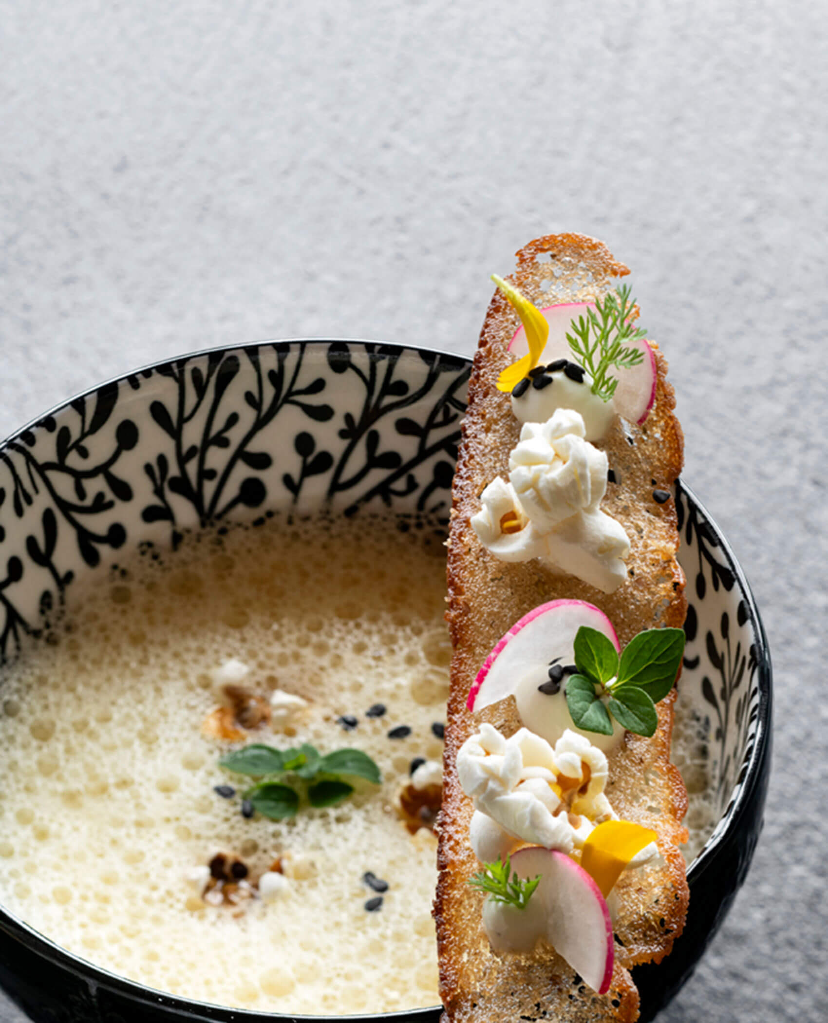 Eine gemusterte Schale mit schaumiger Suppe, die mit Popcorn, Radieschen, Kräutern und Samen dekorativ gekrönt ist. - Hotel Magdalenahof