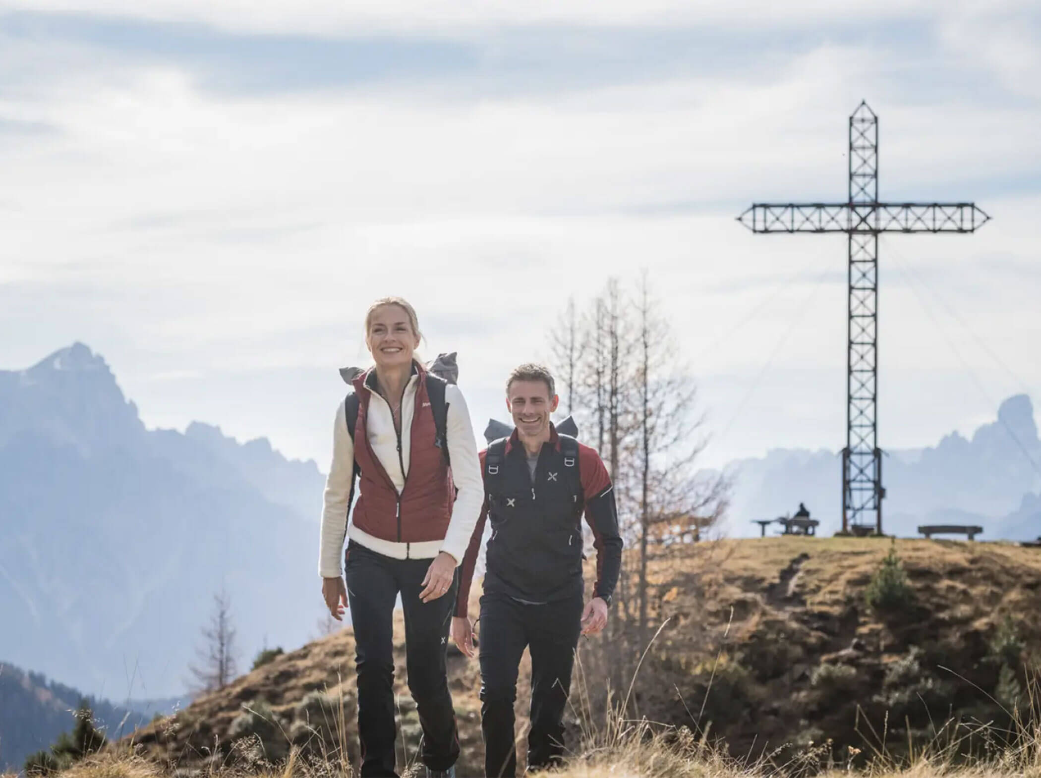 Two people hiking on a grassy hill with a large cross and mountains in the background. - Hotel Magdalenahof