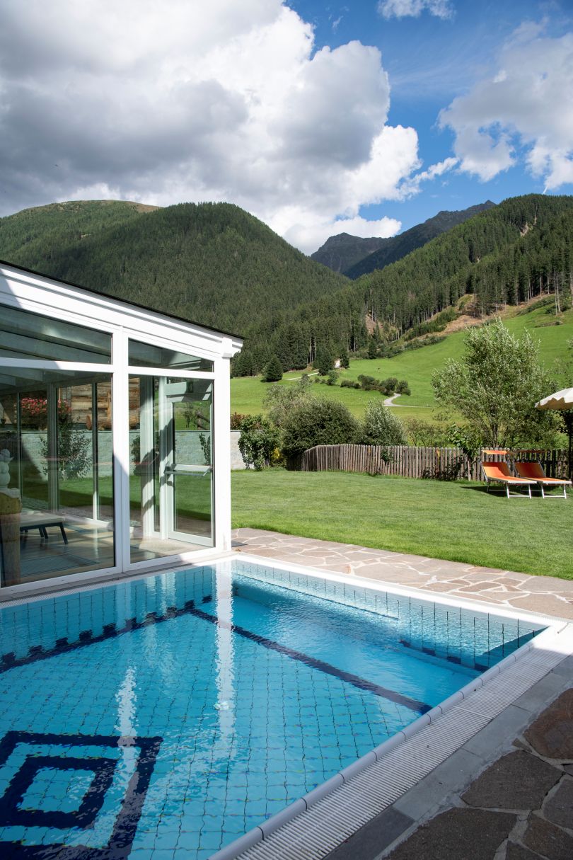 Ein kleiner Pool blickt auf eine Rasenfläche mit Liegestühlen, Berge und Bäume unter einem teilweise bewölkten Himmel. - Hotel Magdalenahof
