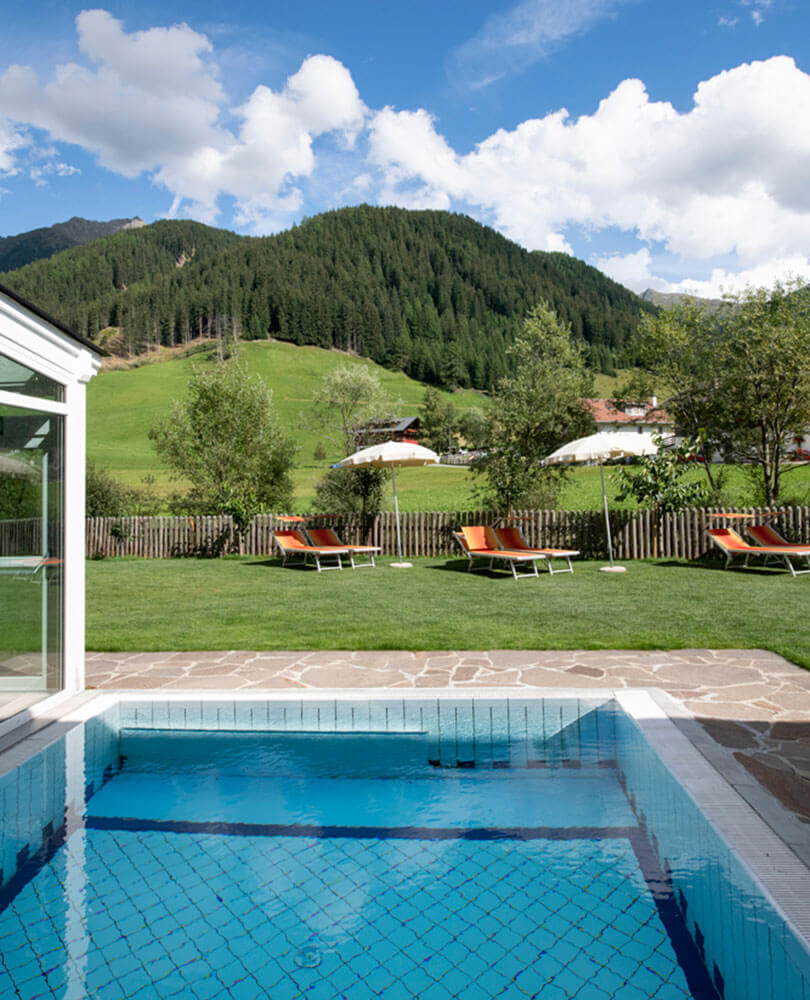 Ein kleiner Pool blickt auf eine Rasenfläche mit Liegestühlen, Berge und Bäume unter einem teilweise bewölkten Himmel. - Hotel Magdalenahof