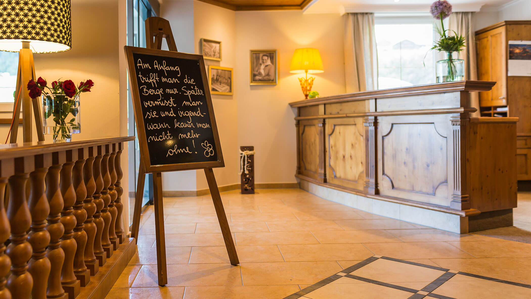 Eine Hotellobby mit einer hölzernen Rezeption, einer Kreidetafel, Lampen und gerahmten Fotos an der Wand. - Hotel Magdalenahof