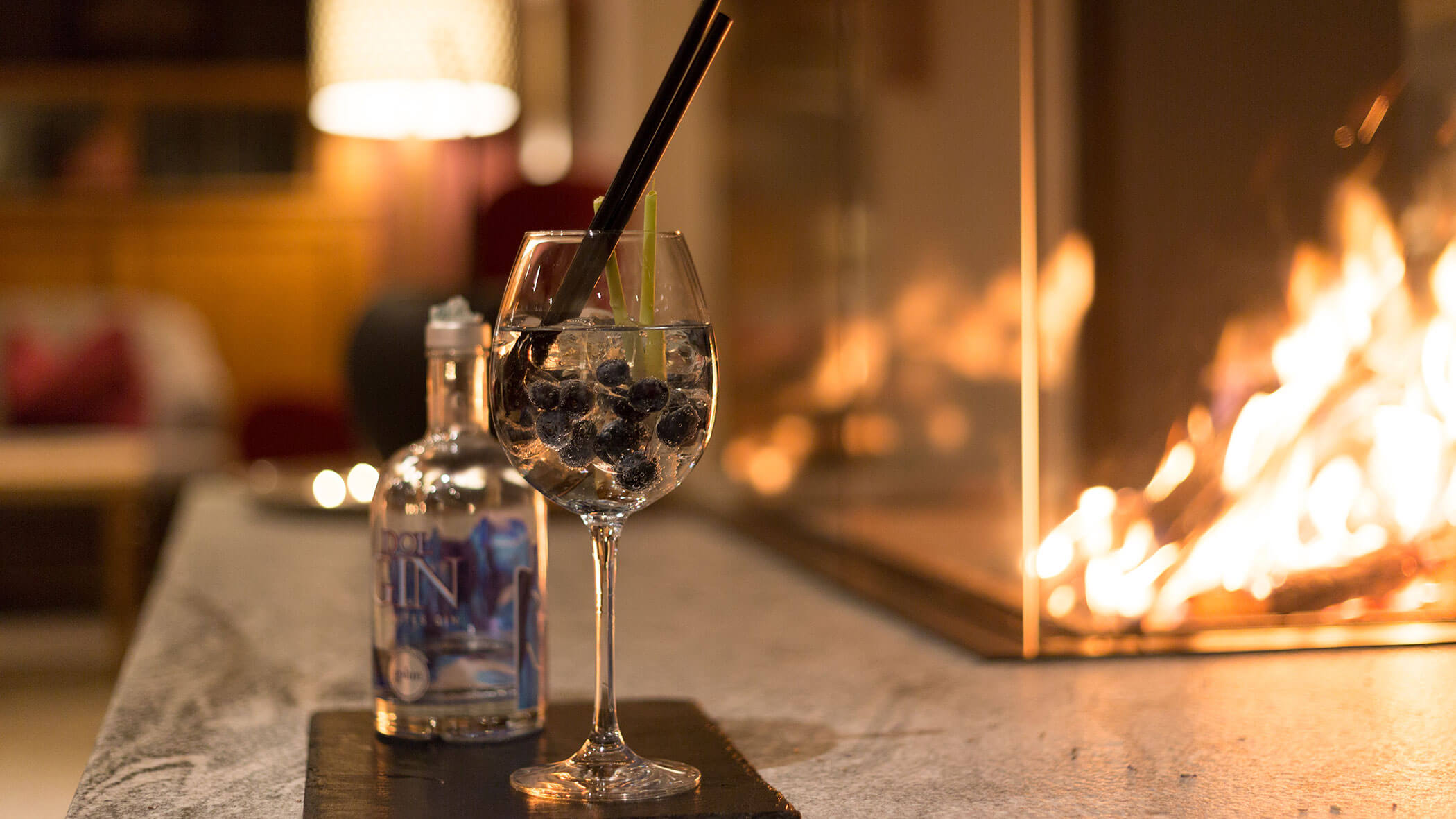 Ein Gin-Cocktail mit Beeren und zwei Strohhalmen steht am Kamin, daneben eine Gin-Flasche. - Hotel Magdalenahof