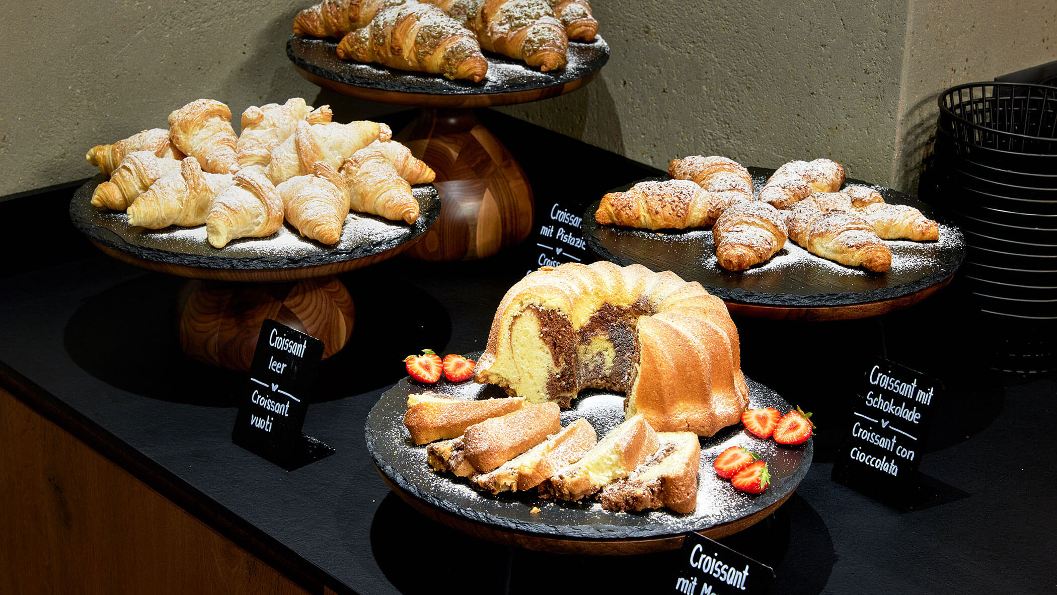 Drei Tabletts mit Croissants und ein Gugelhupf mit aufgeschnittenen Erdbeeren stehen auf einer schwarzen Theke. - Hotel Magdalenahof