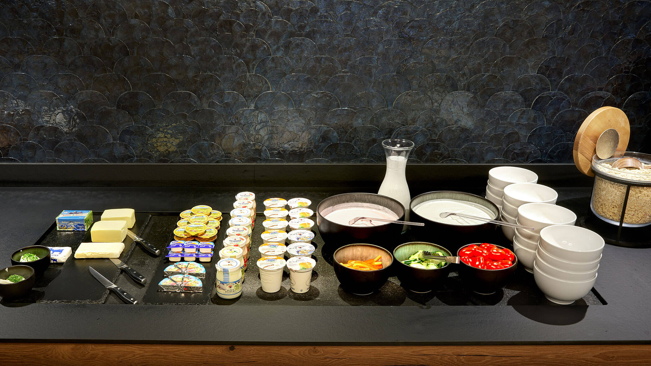 Buffet mit Joghurtbechern, Butter, Käse, Milch, Obst, Gemüse, Schüsseln und einem Behälter mit Müsli. - Hotel Magdalenahof