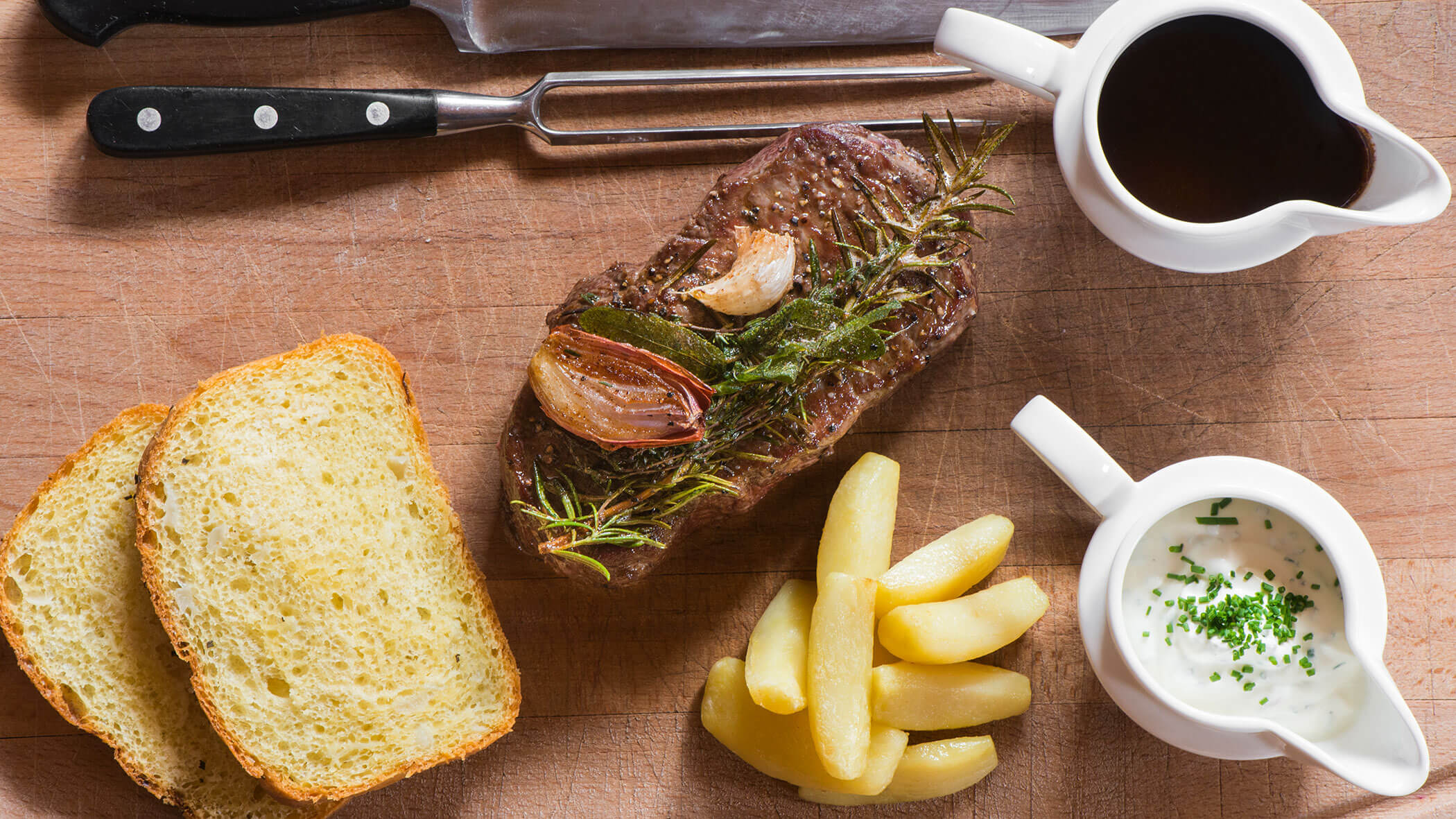 Gegrilltes Steak mit Kräutern und Knoblauch, Brotscheiben, Kartoffelspalten und Soßen auf einem Holzbrett. - Hotel Magdalenahof
