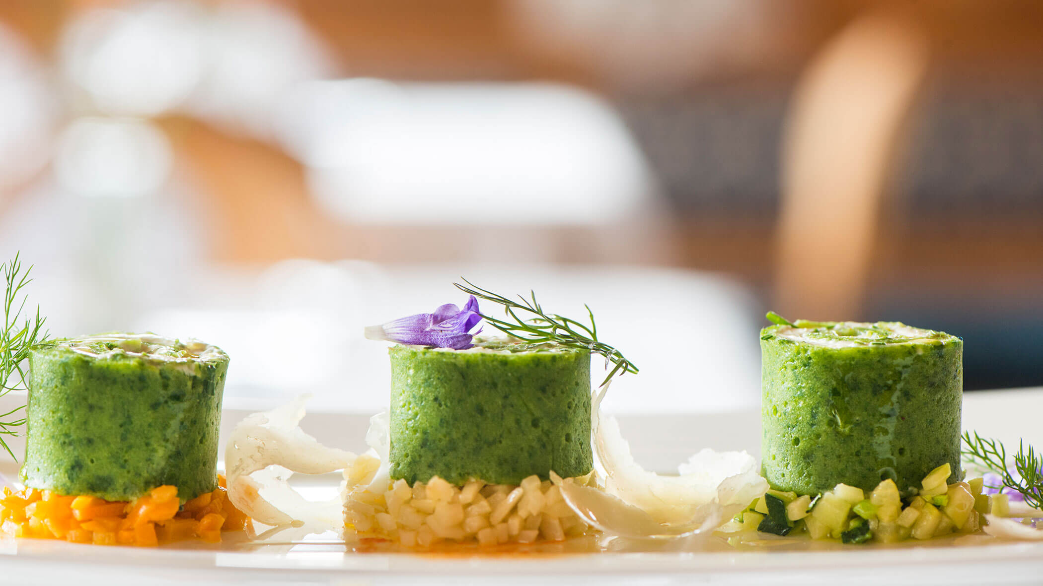 Drei zylindrische grüne Gemüsemousses auf gewürfeltem Gemüse, garniert mit Kräutern und einer violetten essbaren Blume. - Hotel Magdalenahof