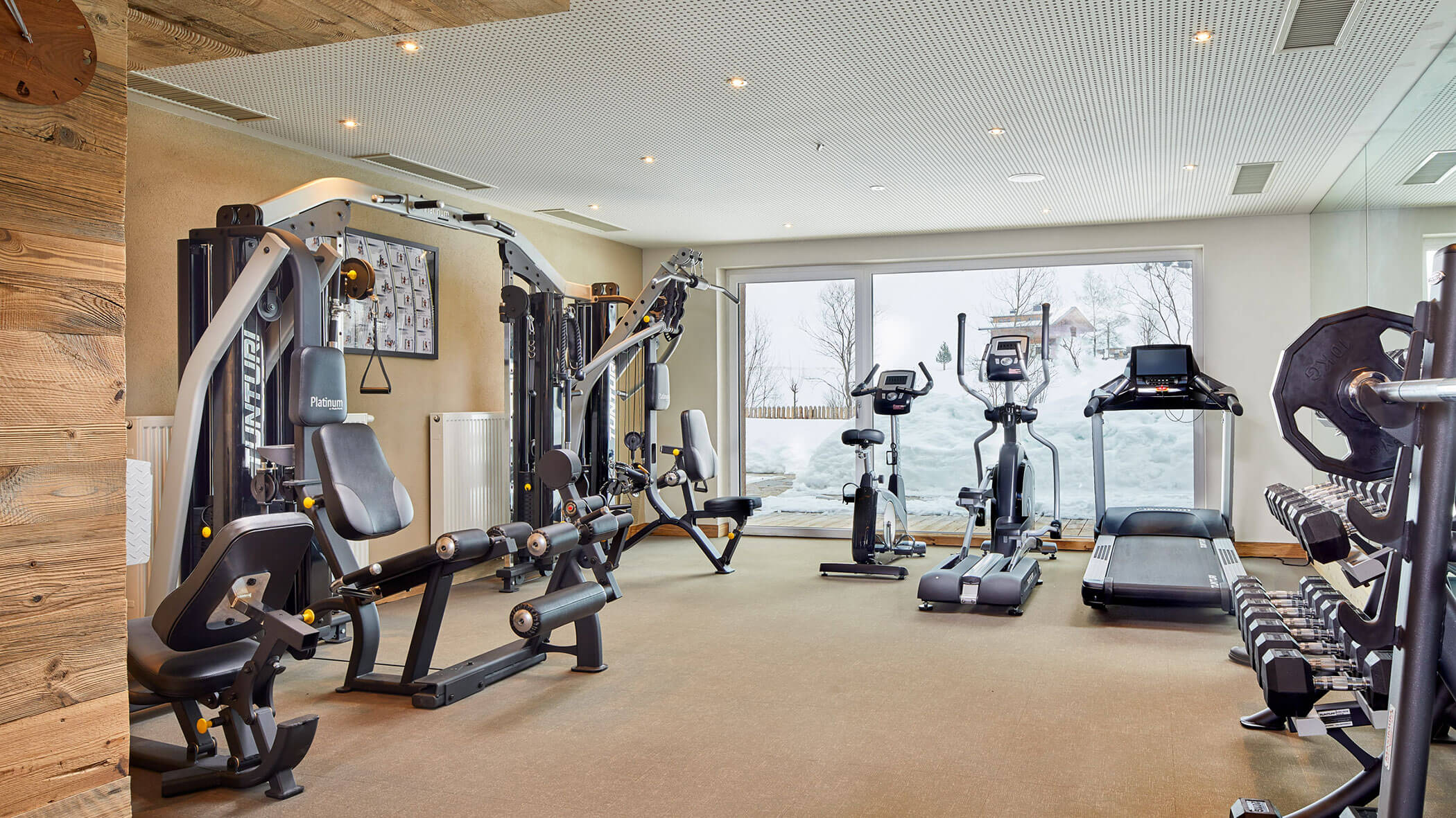 Modernes Fitnessstudio mit verschiedenen Fitnessgeräten, freien Gewichten und großen Fenstern mit Blick auf die verschneite Landschaft draußen. - Hotel Magdalenahof