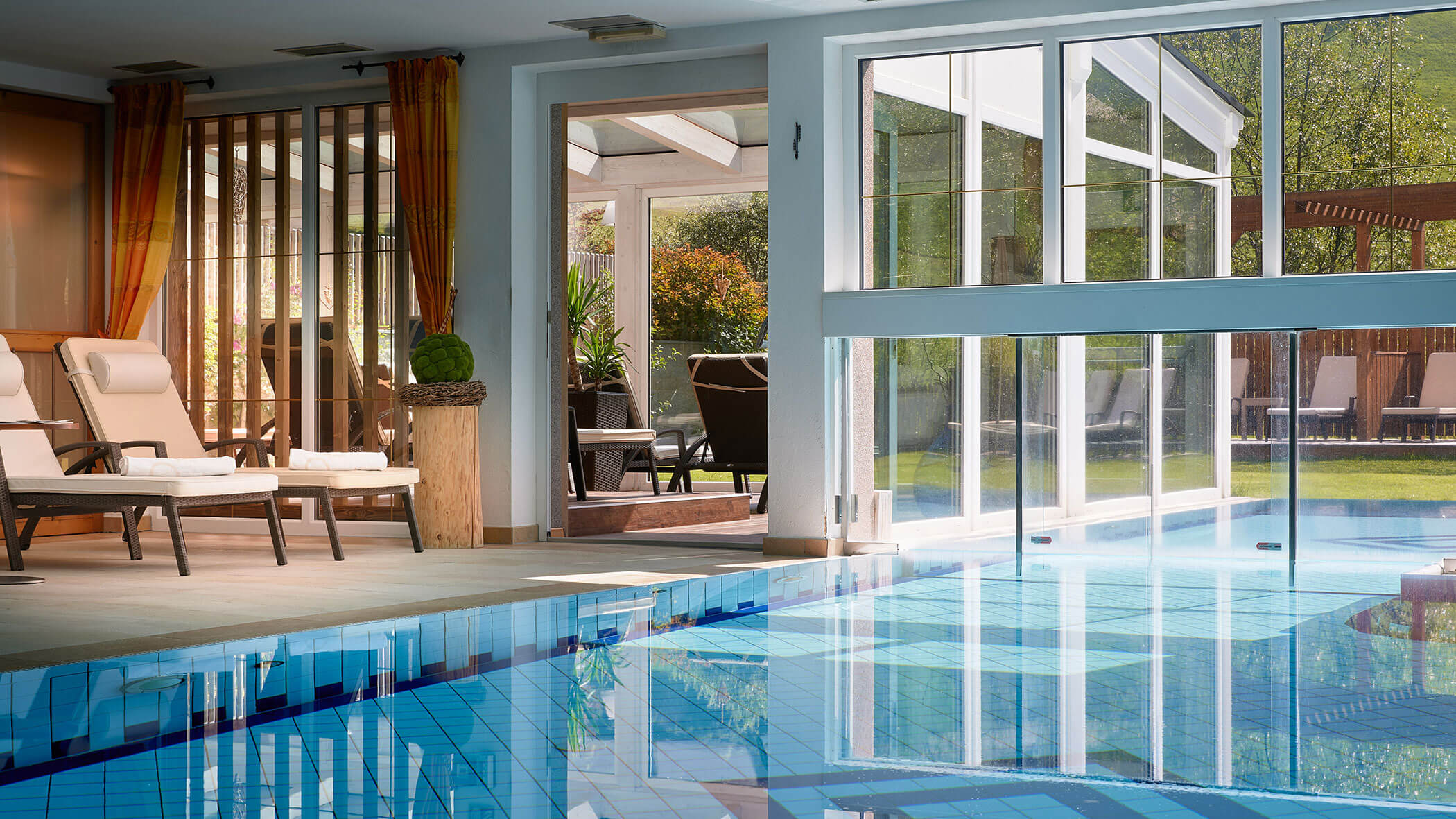 Innenpool mit Liegestühlen neben großen Fenstern und offenen Türen, die zu einer Gartenterrasse führen. - Hotel Magdalenahof