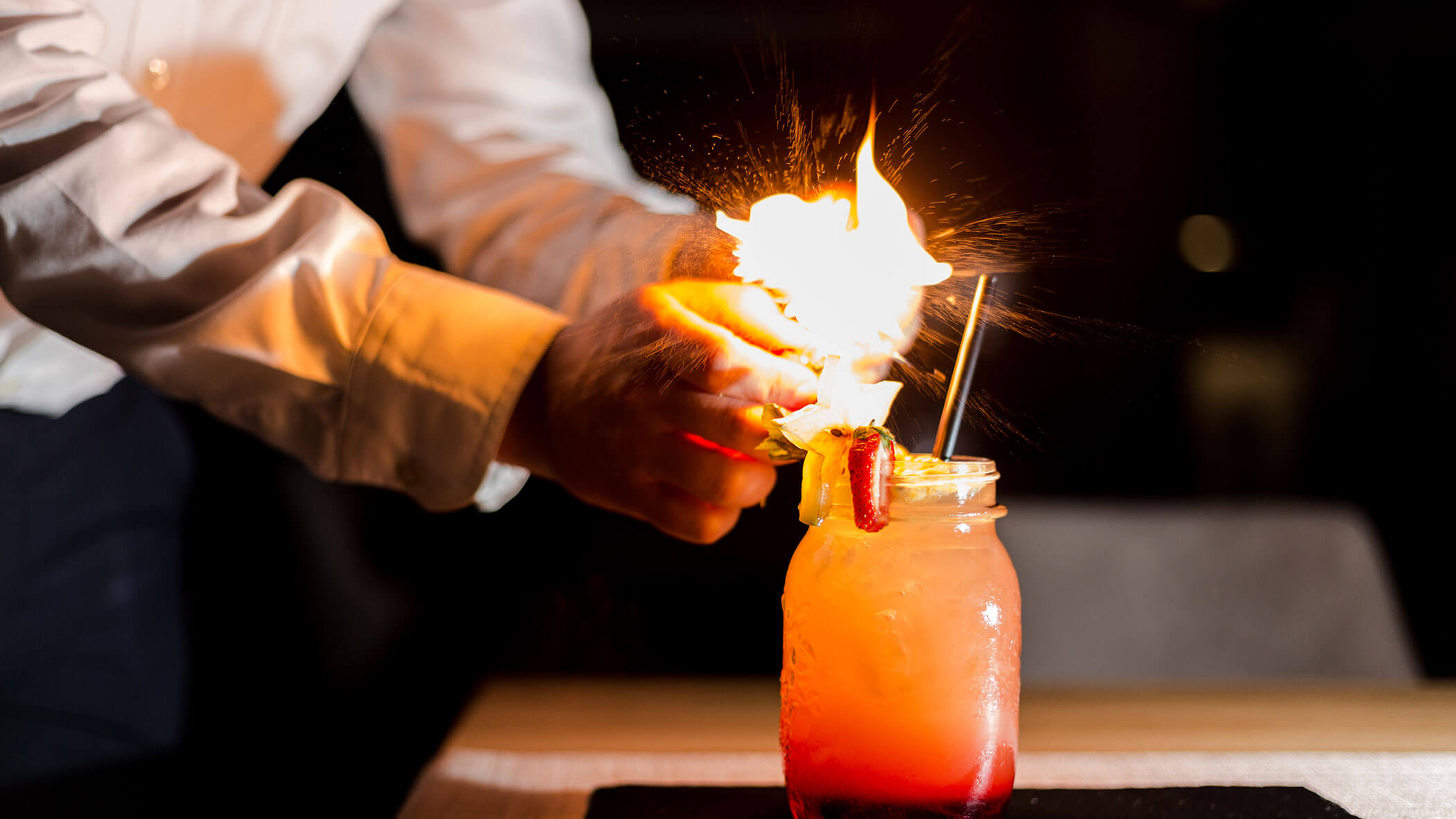 Ein Barkeeper entzündet eine Flamme über einem bunten Cocktail in einem mit Früchten garnierten Einmachglas. - Hotel Magdalenahof